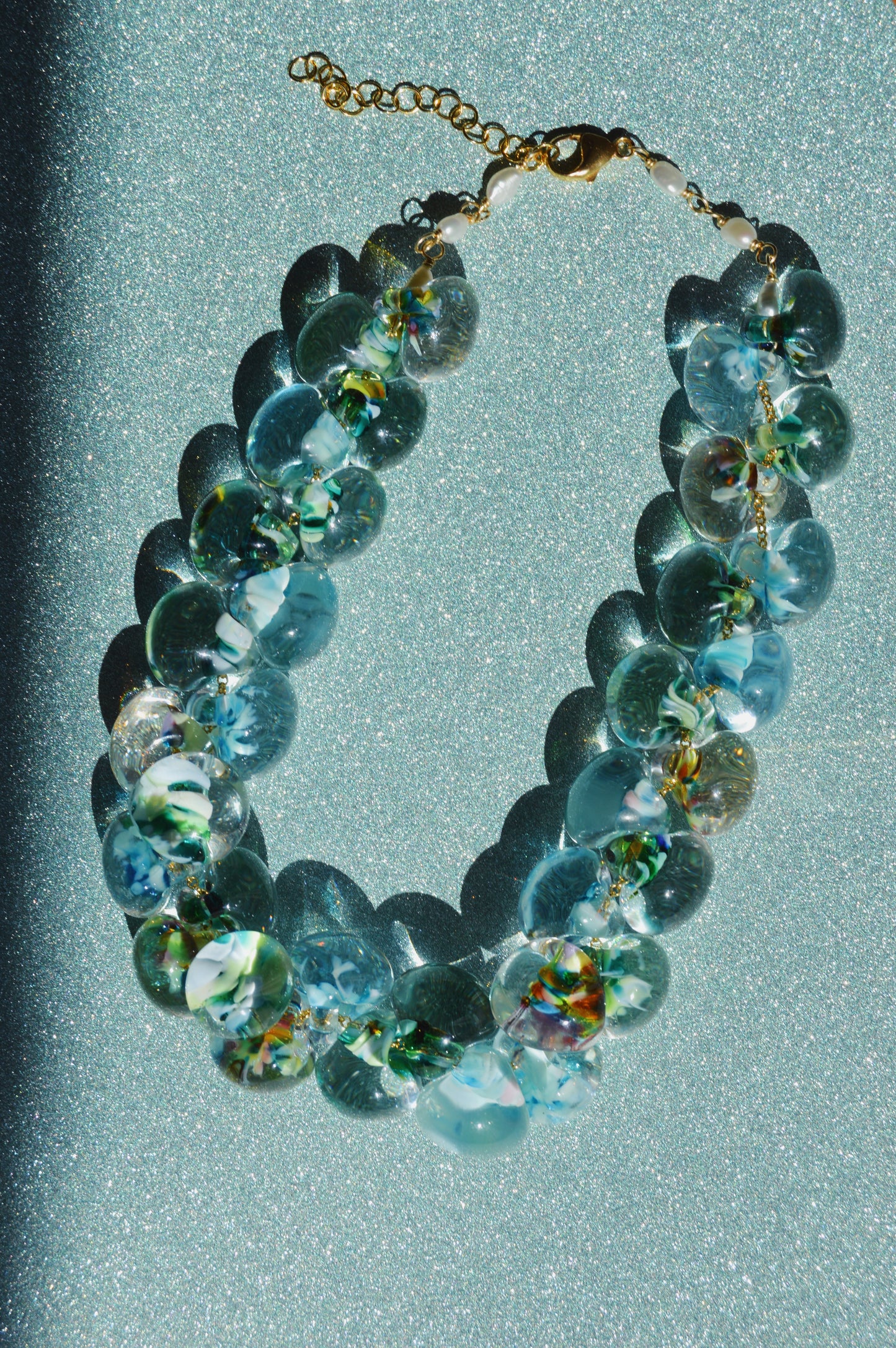 The Grand Bubble Glass Necklace-Tahiti, featuring large, round translucent blue-green glass beads and a gold clasp, is displayed on a glittery light blue background with bold shadows.
