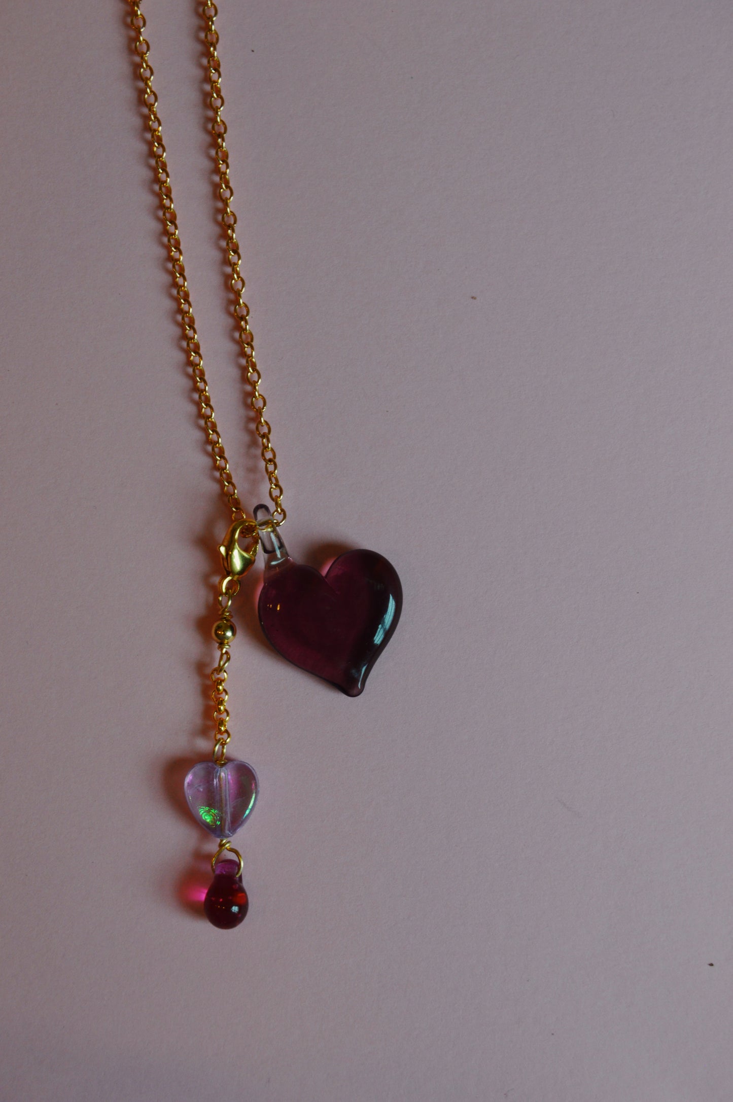 The Sweetheart Charm necklace features a gold chain with three dangling pendants: a large purple glass heart, a smaller clear heart with pink accents, and a tiny round pink bead, shown on a pale pink background.