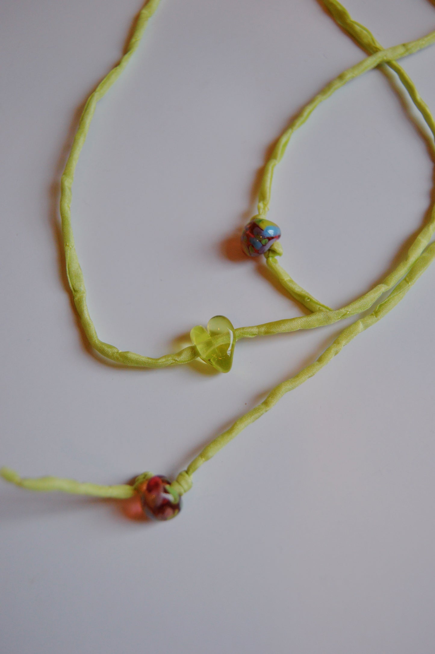 The Mini Silk Glass Heart- Light Green is a yellow fabric artisan necklace with a green handmade lampwork glass heart center and two round multicolored beads, shown on a white background.