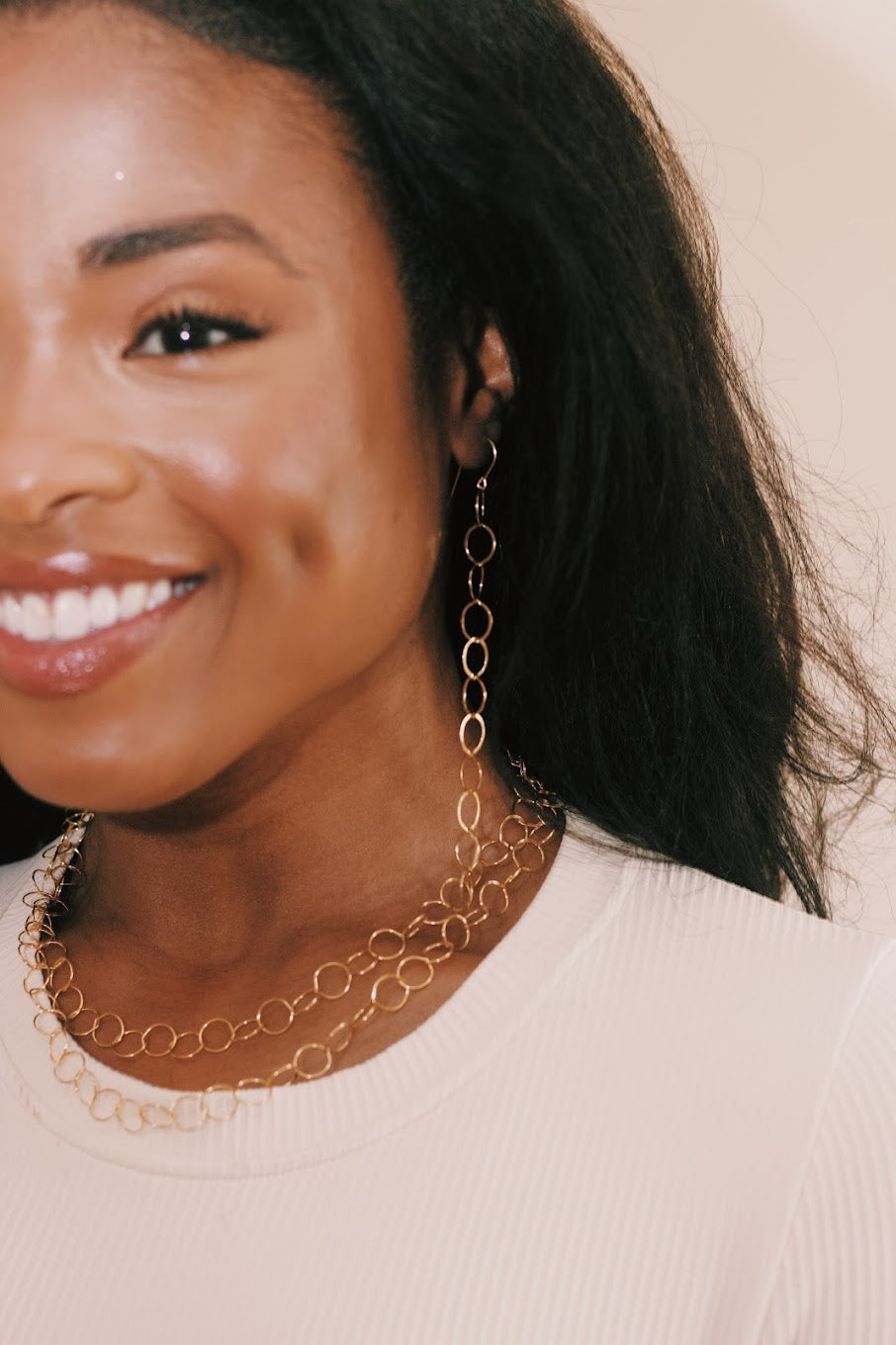 A woman smiles, showing dimples, and wears a cream top with matching 14k gold filled chain earrings and the Gold Filled Circle Chain Necklace. Only the left side of her face and upper body are visible.