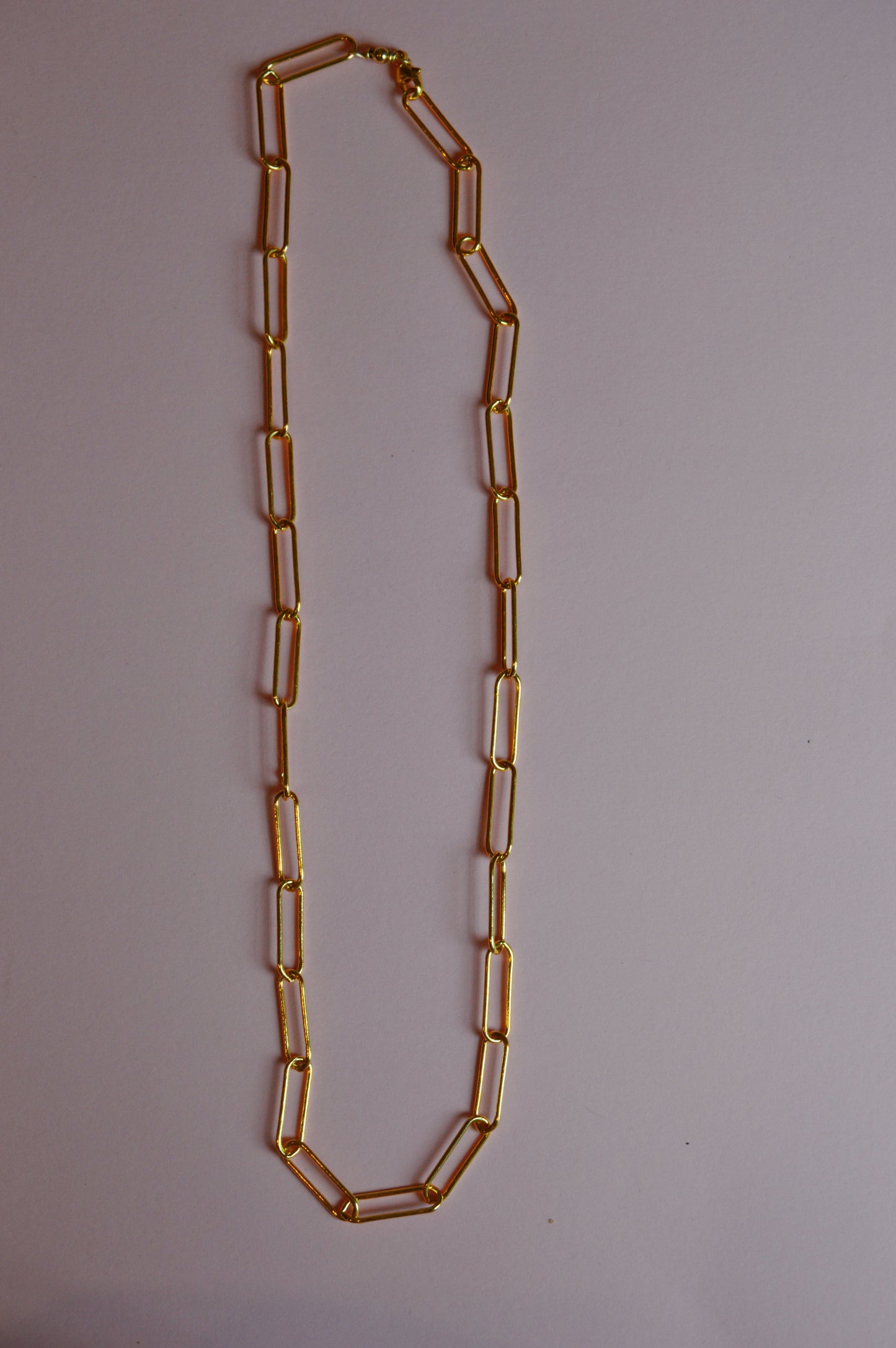 The Paperclip Layer necklace, featuring large elongated rectangular gold links, is displayed on a simple light surface.