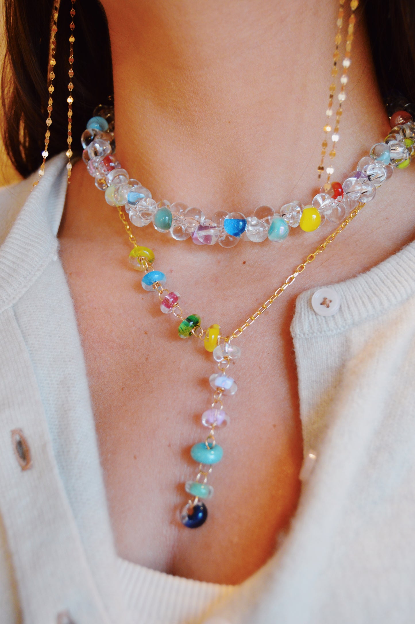 A close-up of someone wearing the Valentine Donut Lariat, a colorful beaded necklace with vibrant, clear beads in various hues, layered over a light buttoned shirt or cardigan.