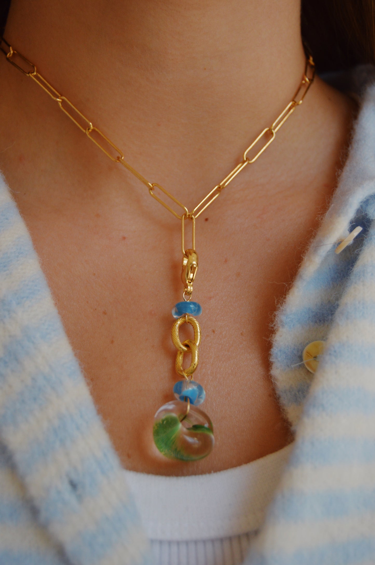 A close-up of a person wearing the Grand Bubble Charm- Green gold chain necklace with blue accents and a clear orb pendant featuring green and blue swirls, accented by lampwork glass beads, over a white top and blue-striped shirt.