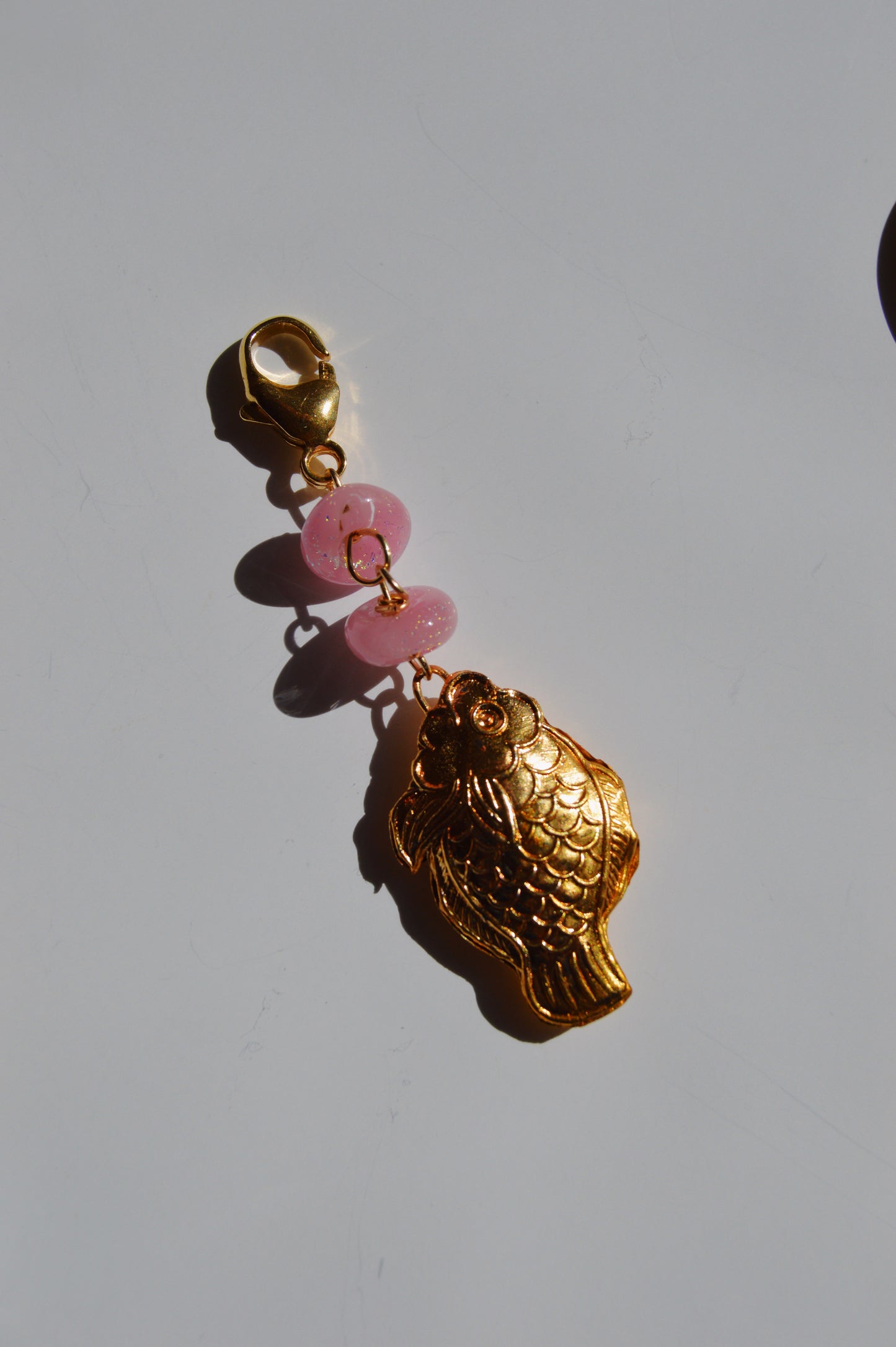The Glass and Fish Charm features a gold cloisonné fish with a clasp, accented by two handmade lampwork glass beads, displayed on a white background.
