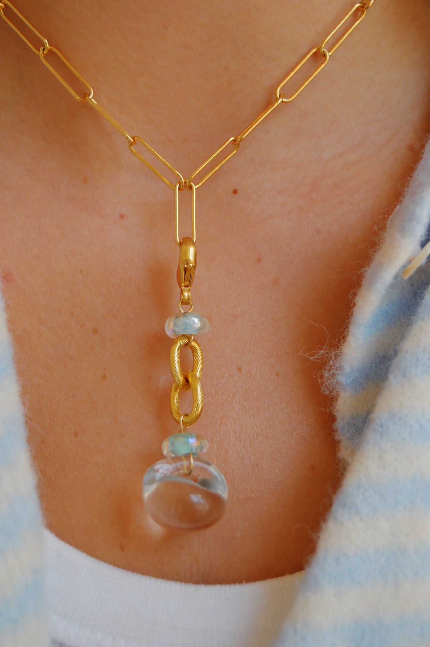 A close-up of the Grand Bubble Charm- Light Blue: a 14k gold-filled chain necklace with a clear round stone pendant, linked gold ring, small opal beads, and handmade lampwork glass beads, worn with a blue and white garment.