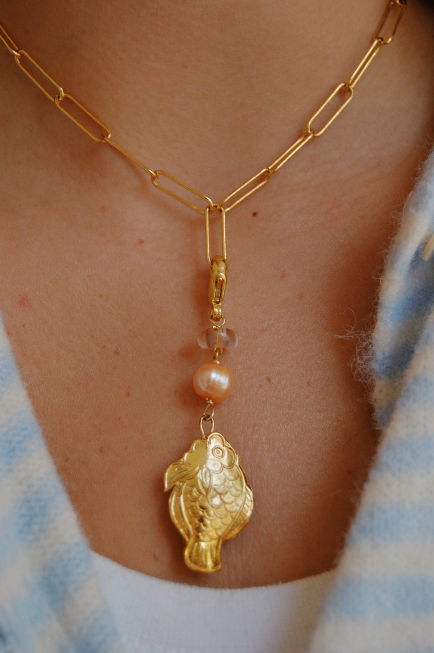 Wearing a Fish and Pearl Charm, this gold paperclip chain necklace features a dangling cloisonné fish, small bead, and cultured freshwater pearl. It's styled with a white top and blue plaid shirt.