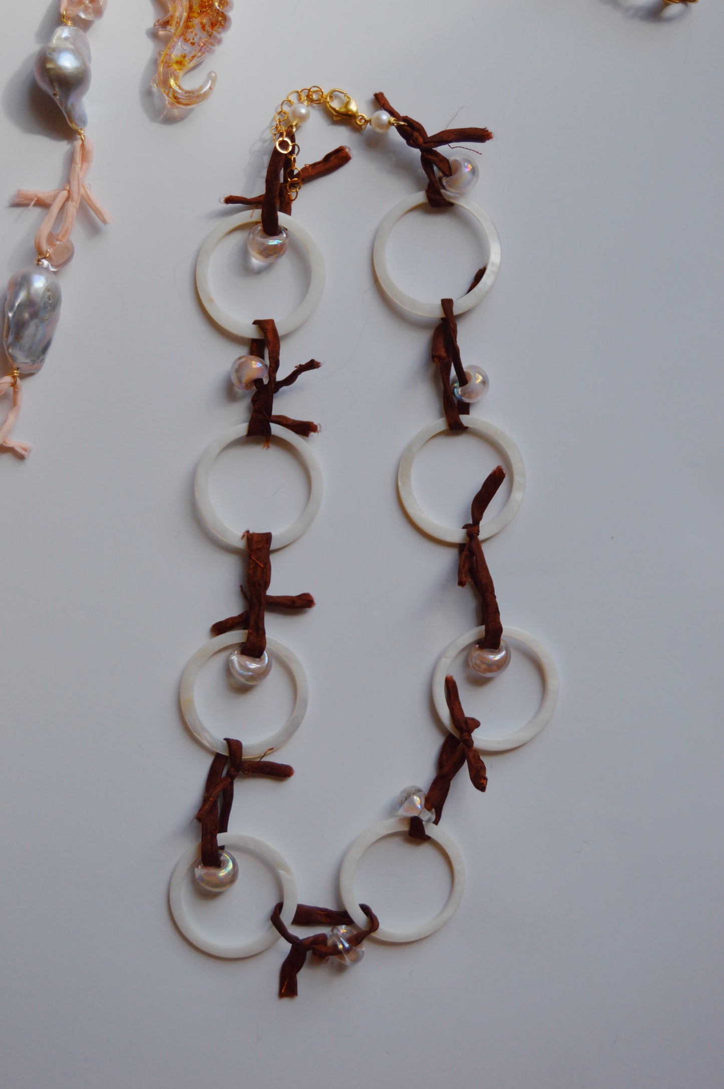 The Silk and Circle Layer is a handmade jewelry piece with large white circular links, brown leather ties, and small pearls, displayed on a white surface with glimpses of other jewelry in the corners.