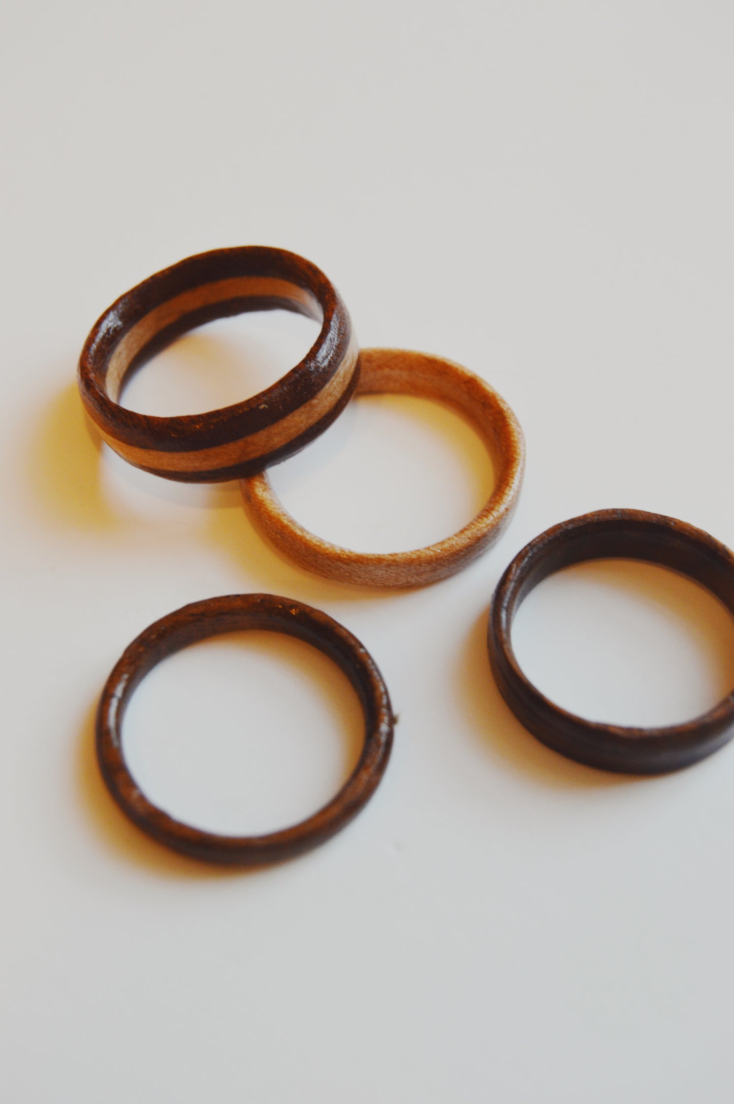 Four artisan-made Wooden Ring- Striped rings in different brown shades, including one from white oak, are displayed on a white surface. Each has a smooth finish and simple design, with one ring lighter than the rest.