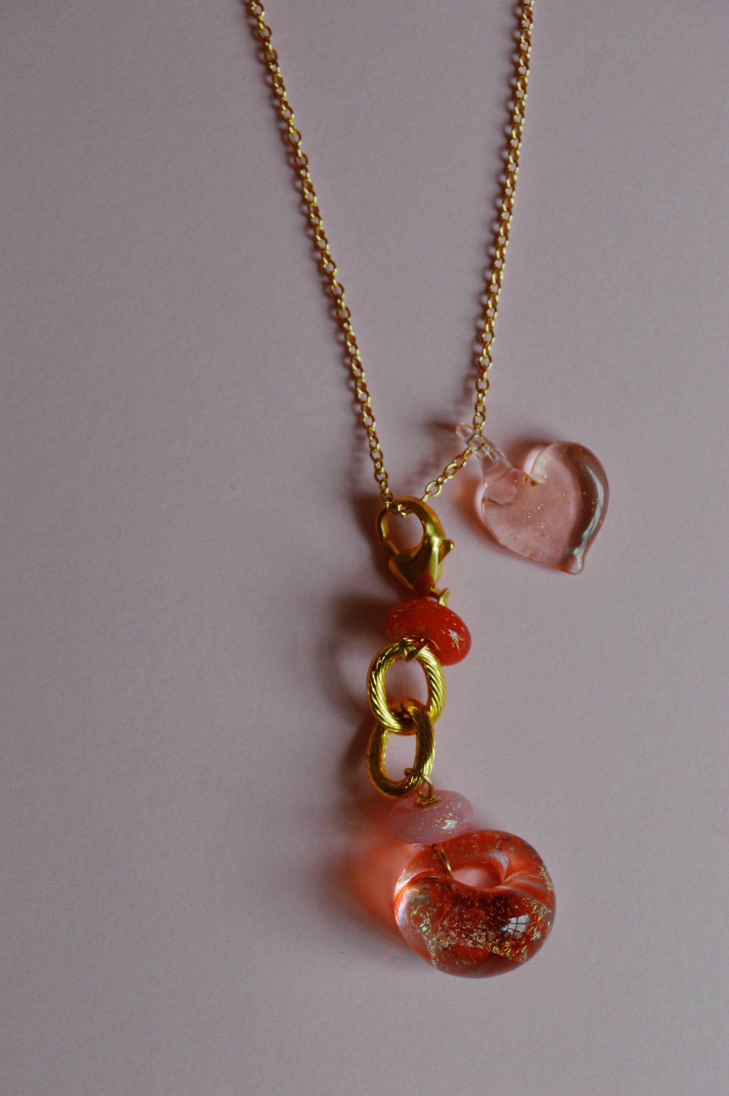 The Valentine Grand Bubble Charm is a gold chain necklace featuring a pink glass heart, gold links, and a circular pink and clear glass pendant with glitter accents, shown on a light pink background.