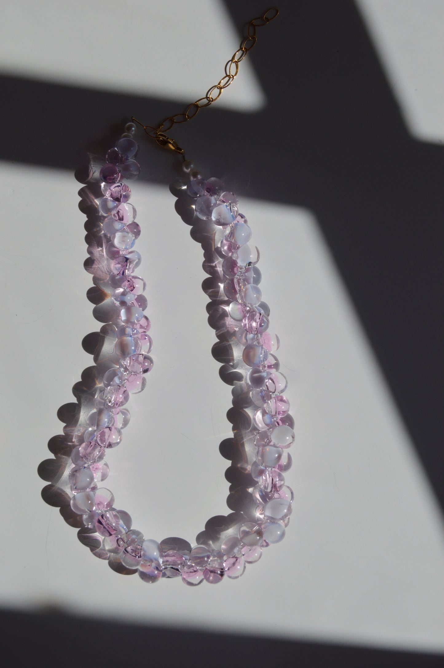 The Bubble Glass Necklace- Pink and Lavender, a double-strand handmade piece featuring translucent pink and lavender beads, is displayed on a white surface with dramatic shadows and sunlight highlighting its gold-filled findings.