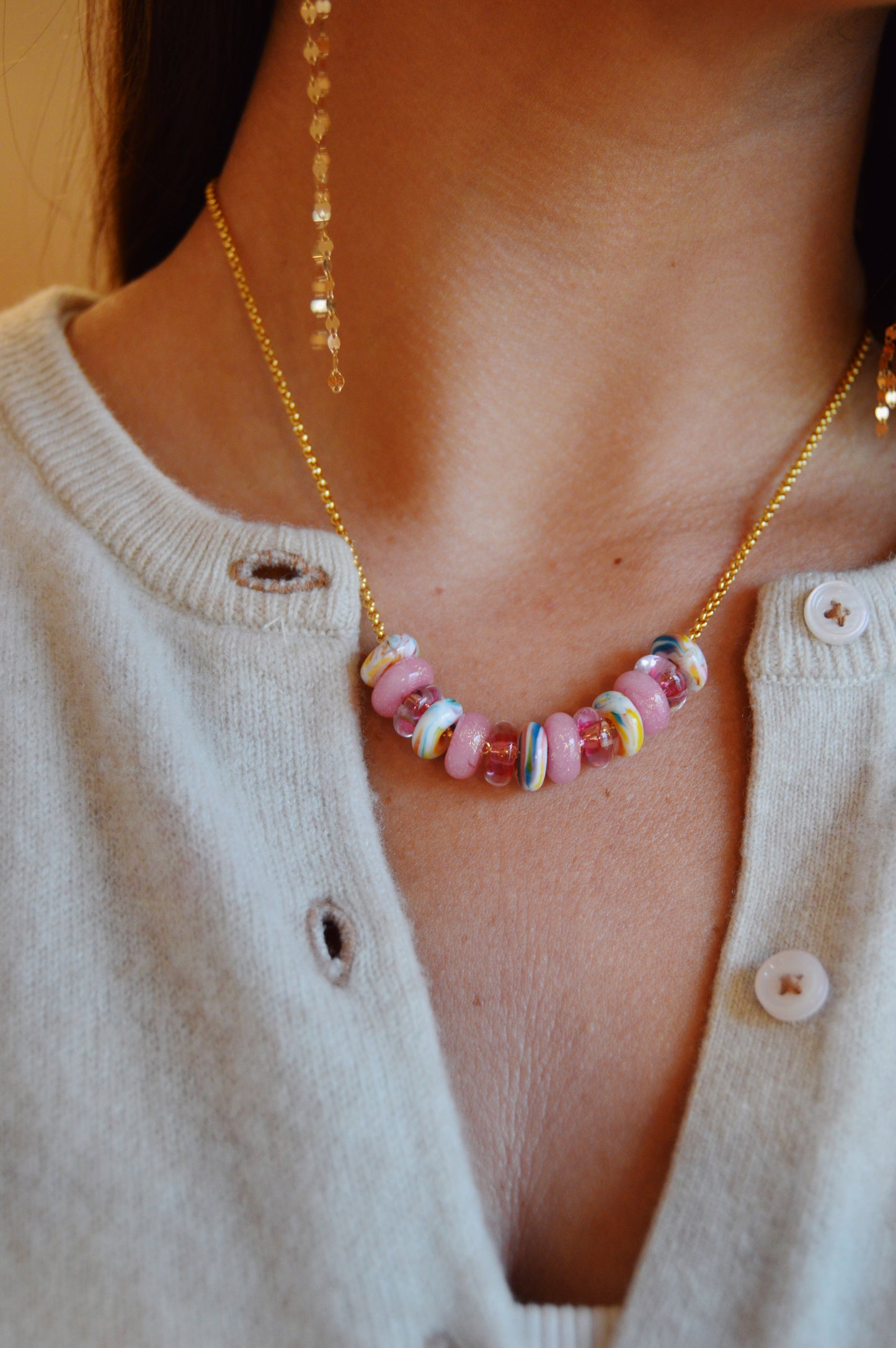 Close-up of a person wearing a beige sweater with white buttons and the Valentine Lots of Donuts Layer—a gold necklace adorned with colorful, round glass beads in pink, blue, yellow, and white. A beaded earring is partially visible.