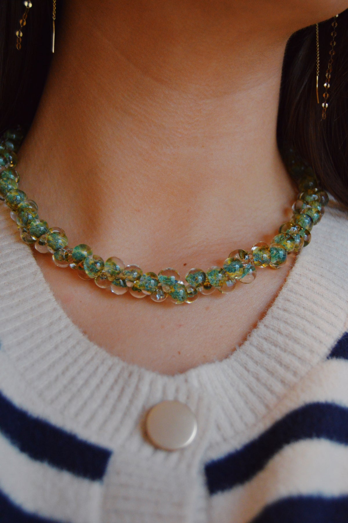 A close-up of someone wearing the Bubble Glass Necklace- Clover, a handmade piece with green and clear glass beads and 14k gold-filled findings, styled with a white and navy striped sweater featuring a silver button.