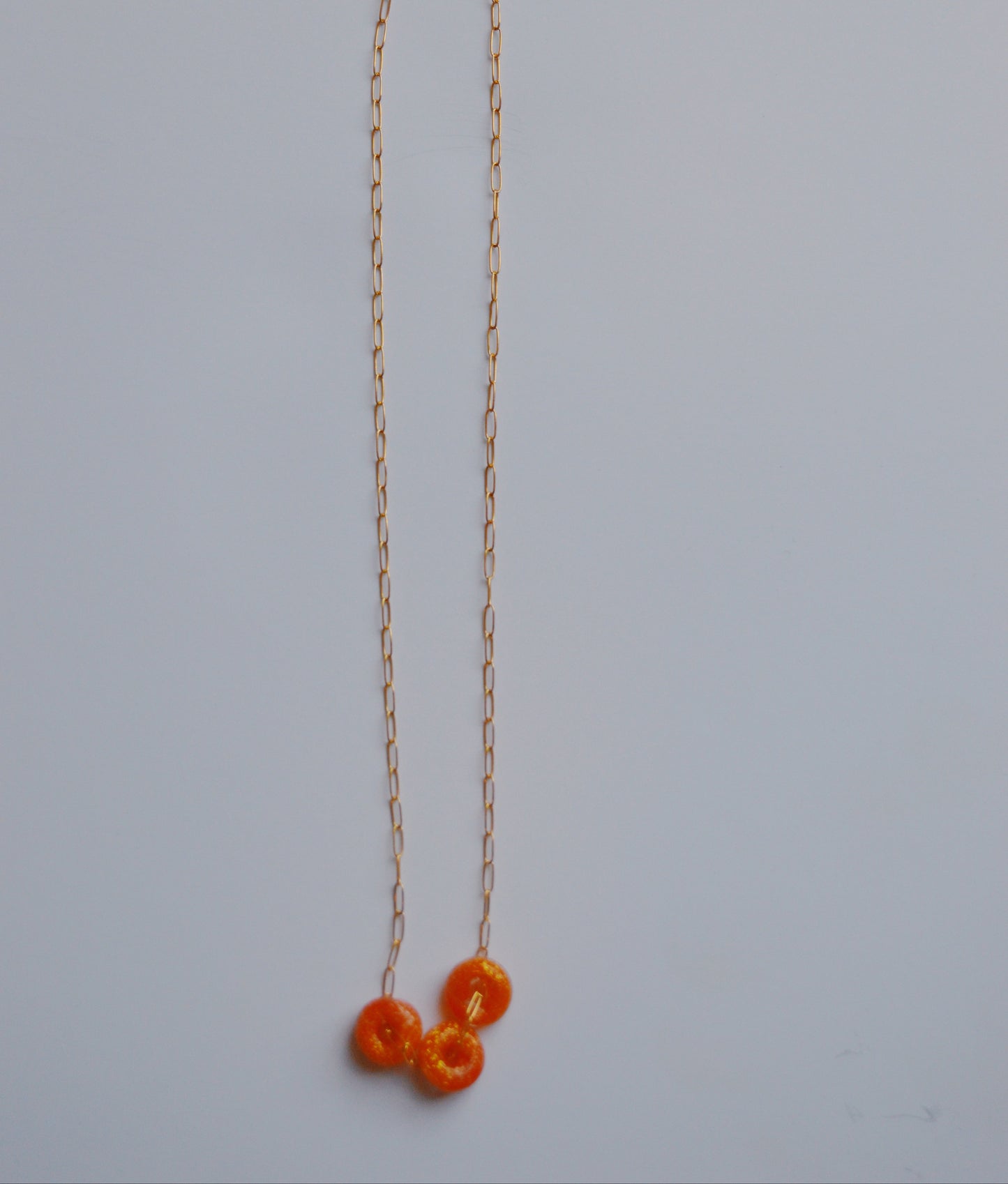 The Dainty Donut Layer- Sunshine features a delicate gold-filled chain with three small orange fuzzy pom-poms at the center, highlighting handcrafted charm against a light gray background.