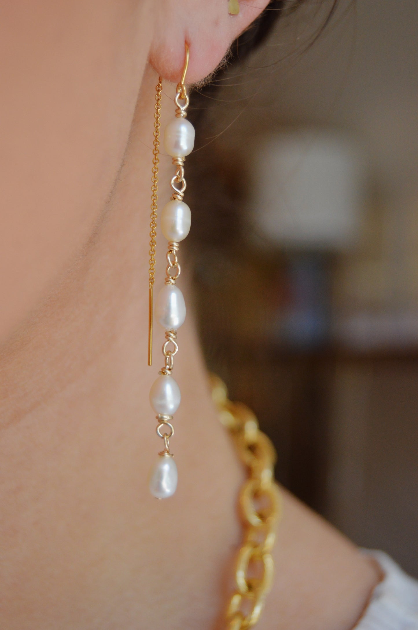 Close-up of a person wearing Pearl Threaders—long 14K gold-filled earrings adorned with five cultured freshwater pearls—set against a softly blurred indoor background.