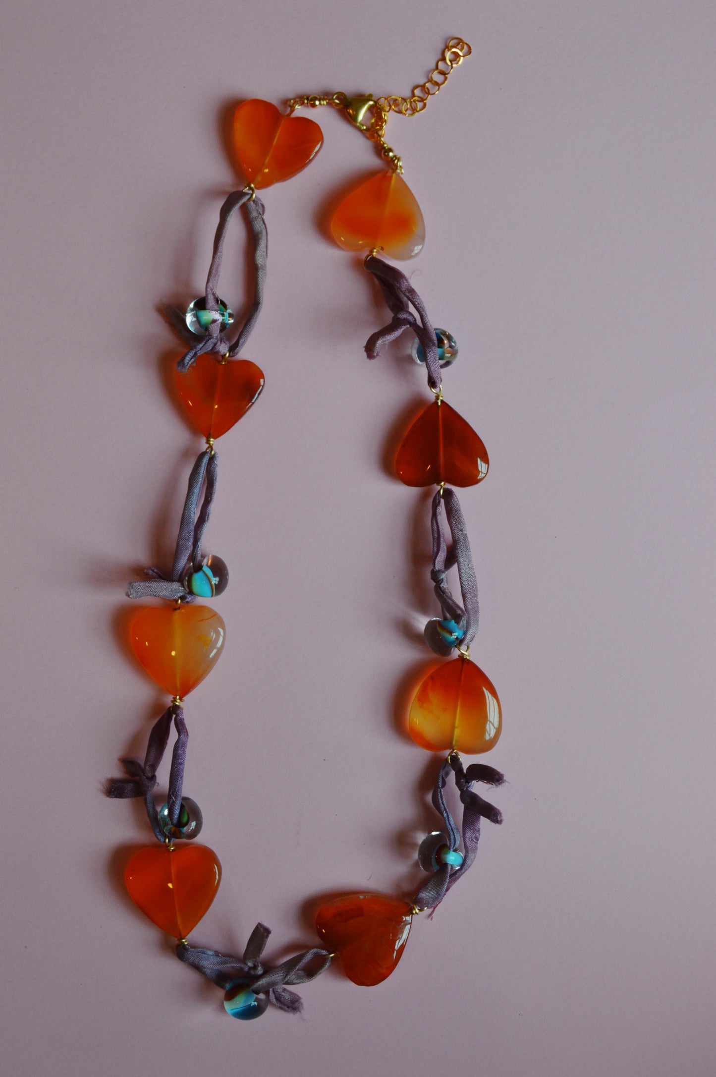 The Silk and Gemstone Heart Layer features large orange heart-shaped beads, blue round beads, and purple ribbon between each bead, displayed on a pale pink background.