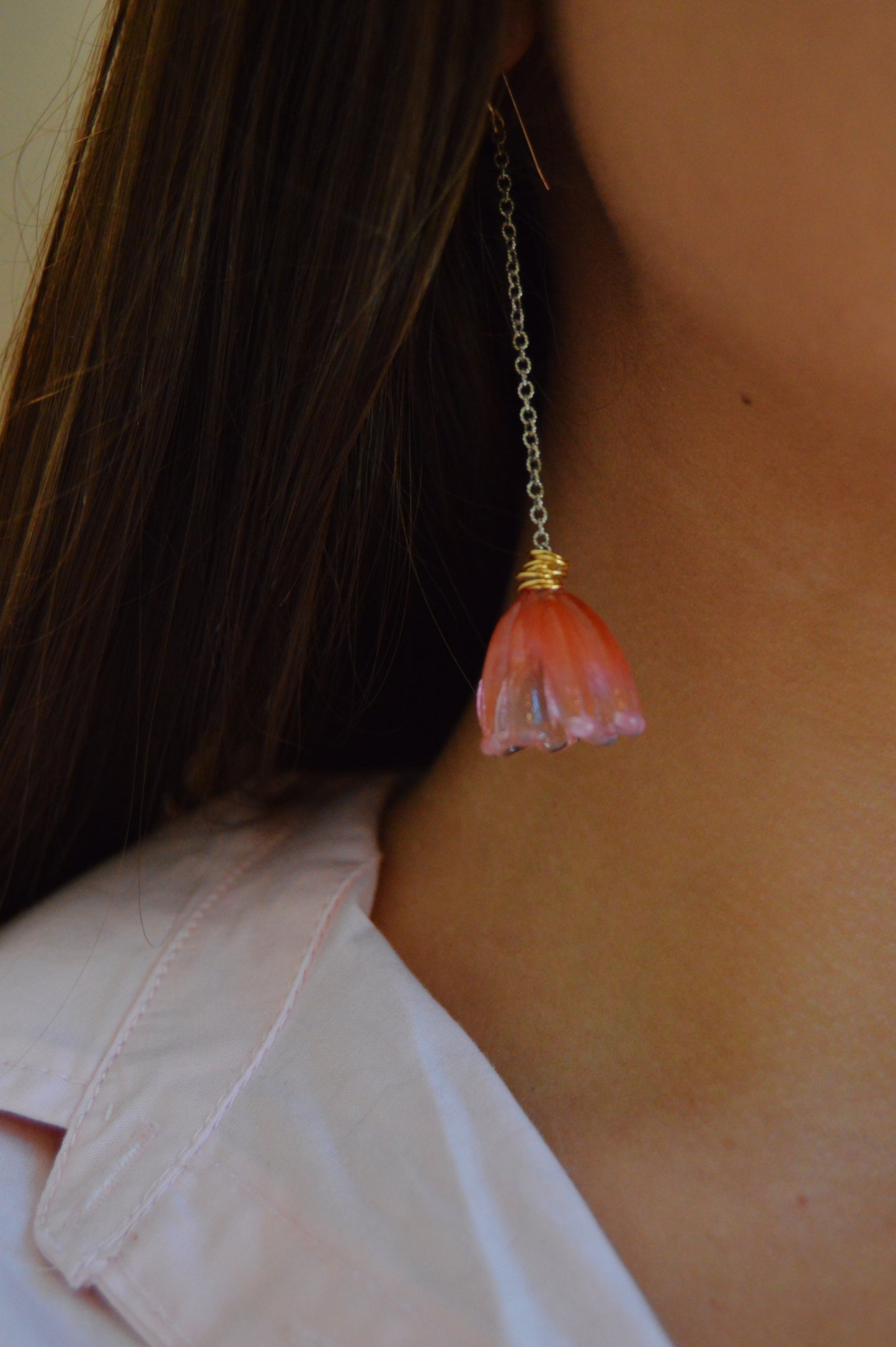 A close-up of a person wearing the Valentine Mixed Metal Tulips earring—a long gold chain style with a pink, flower-shaped pendant. The individual has straight brown hair and is dressed in a light pink collared shirt.