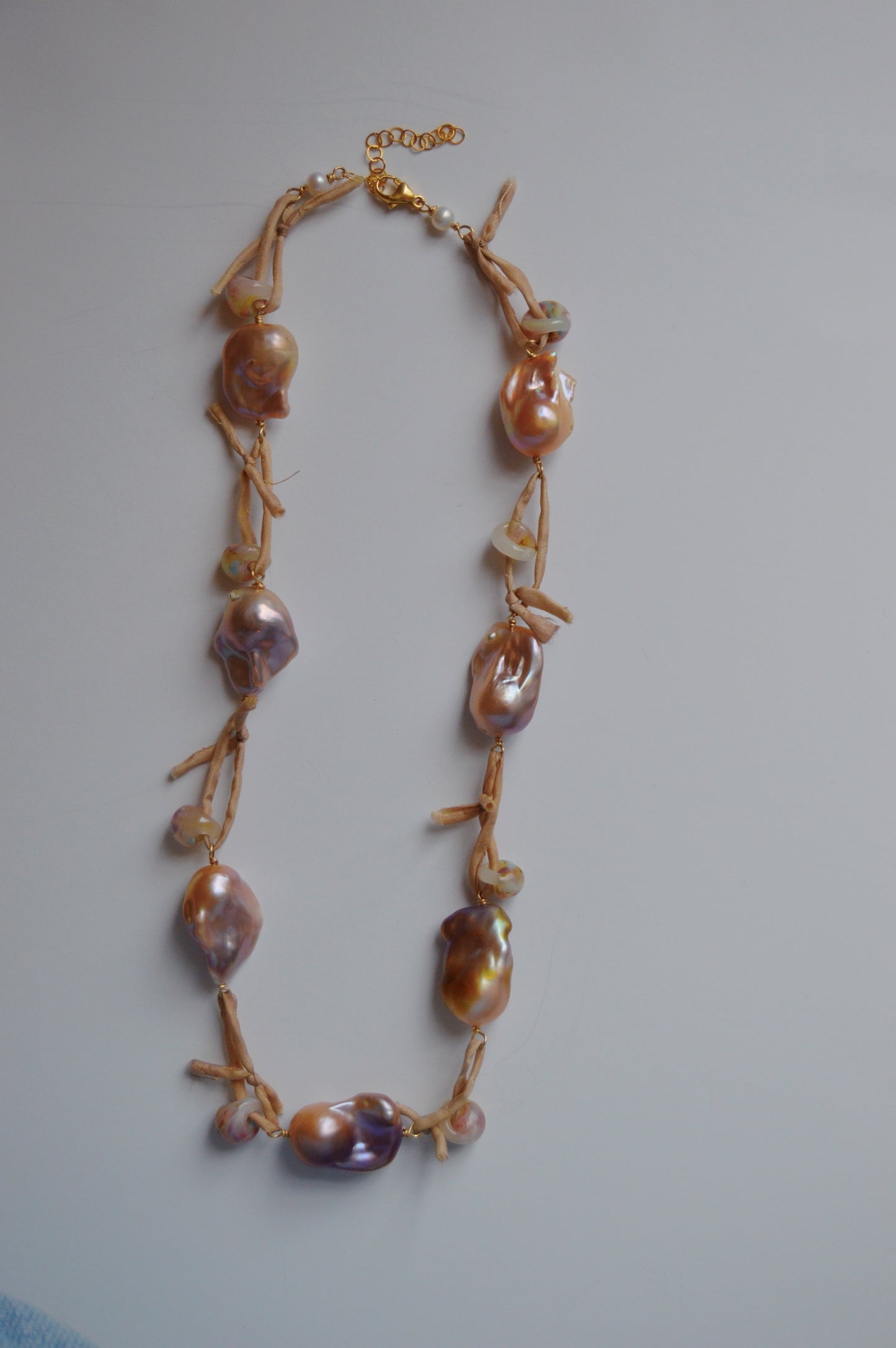 The Silk and Pearl Layer- Sandy Puka necklace showcases large, irregular shell beads on a light brown cord with knots between each bead and a gold clasp, all set against a plain white background.