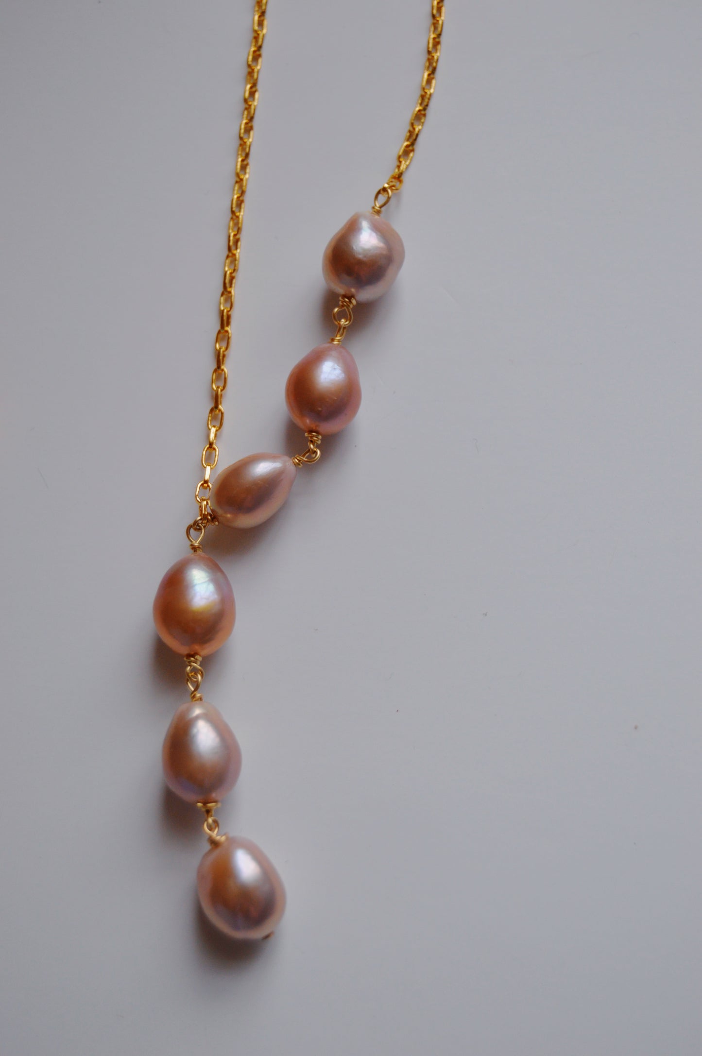 The Champagne Pearl Lariat features six vertically arranged, pink-lavender cultured freshwater pearls on a delicate 14k gold-filled chain—handcrafted for timeless elegance.