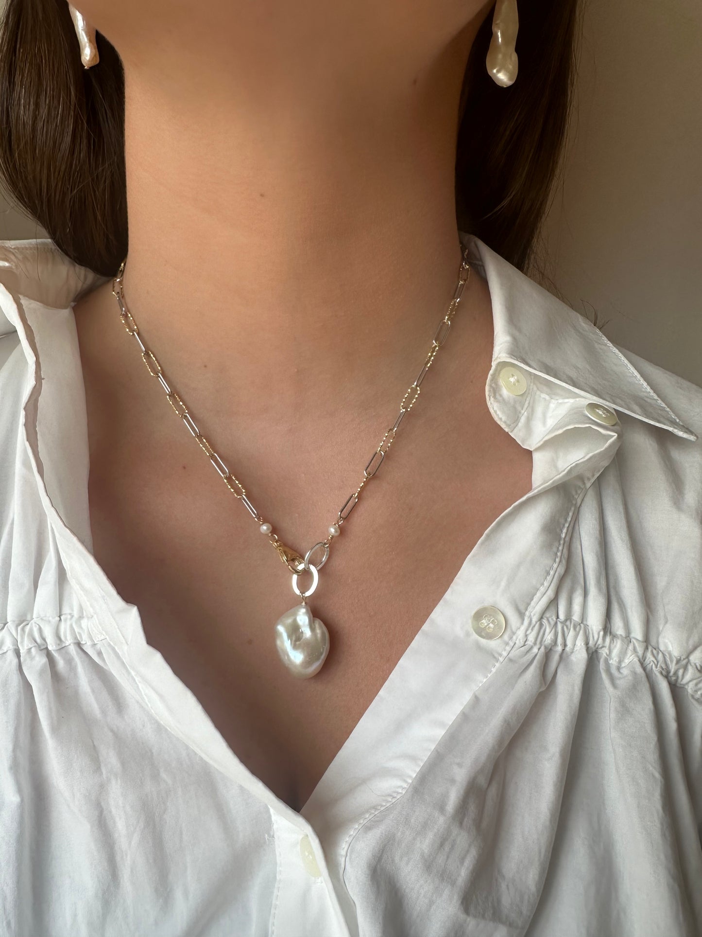 A woman in a white button-up shirt showcases the Showstopper Baroque Pearl necklace, featuring a mixed metal chain with a large baroque pearl pendant and matching pearl drop earrings.