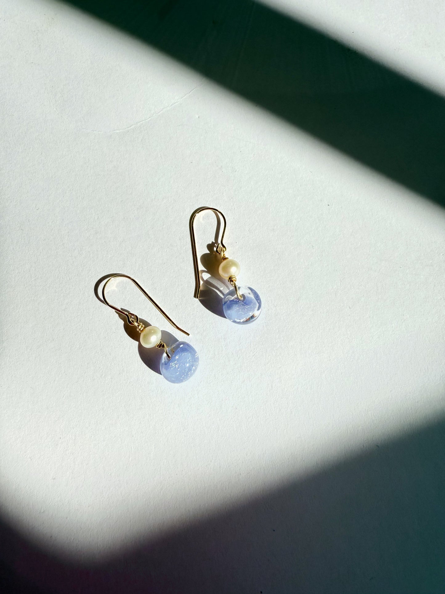 The Bubble Minis, a pair of gold hook earrings with freshwater pearls and round light blue glass beads, rest on a white surface bathed in gentle sunlight and shadows.