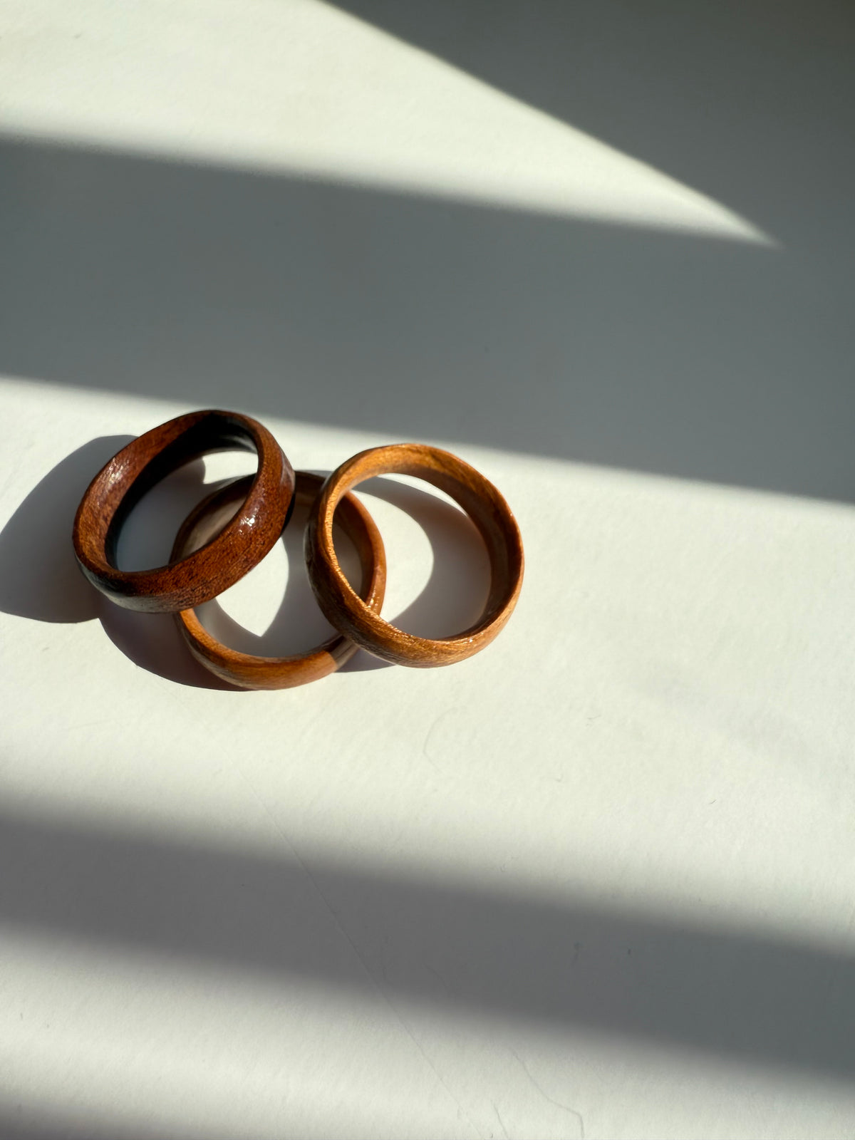 The Wooden Ring- Medium Wood rests on a white surface, bathed in bright sunlight that casts striking shadows and diagonal patterns across the scene.