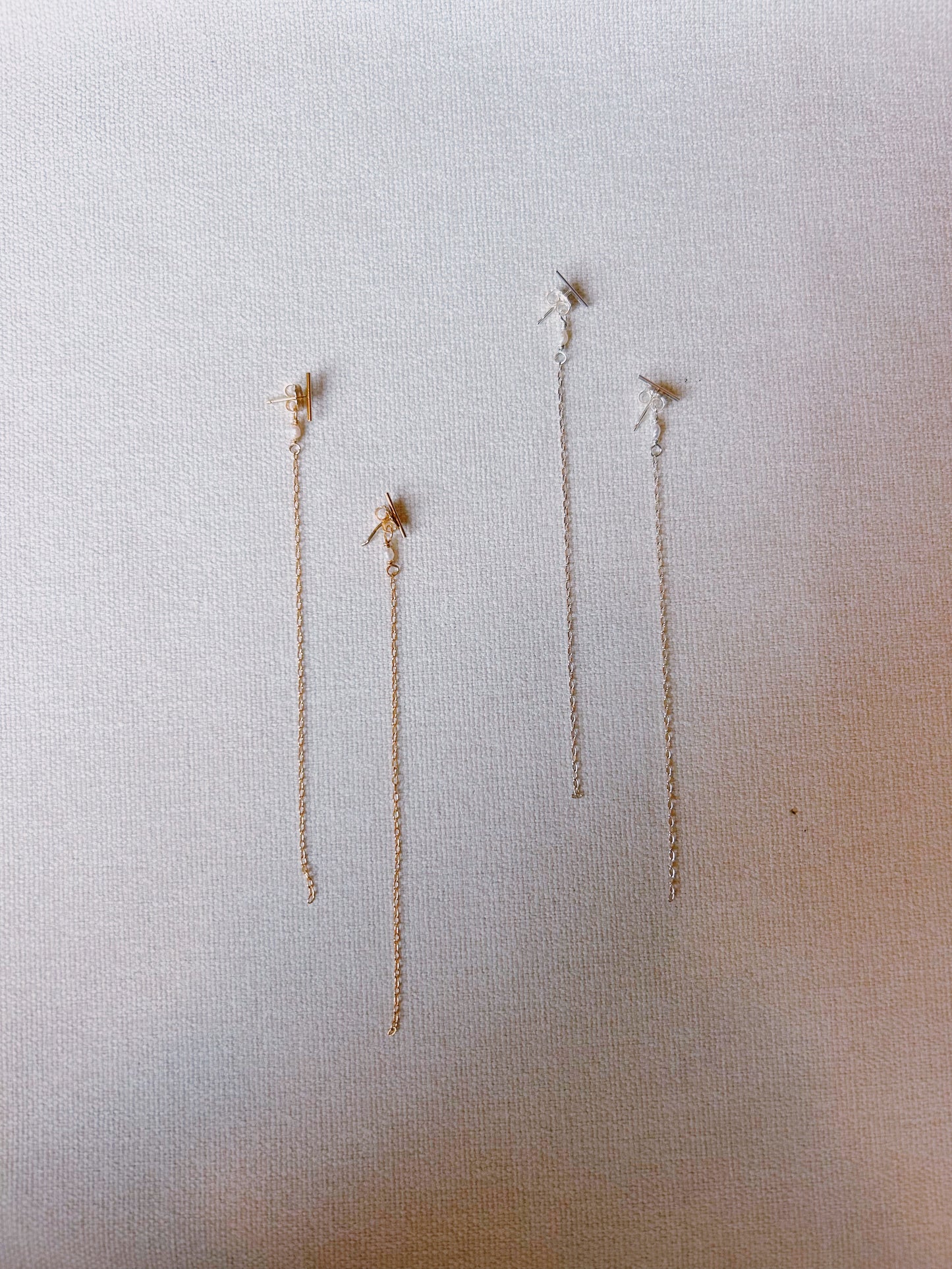 Four KK x Poema 14k gold filled threader earrings, featuring small decorative elements and freshwater pearls, are displayed on a light textured fabric, with two pairs arranged vertically side by side.