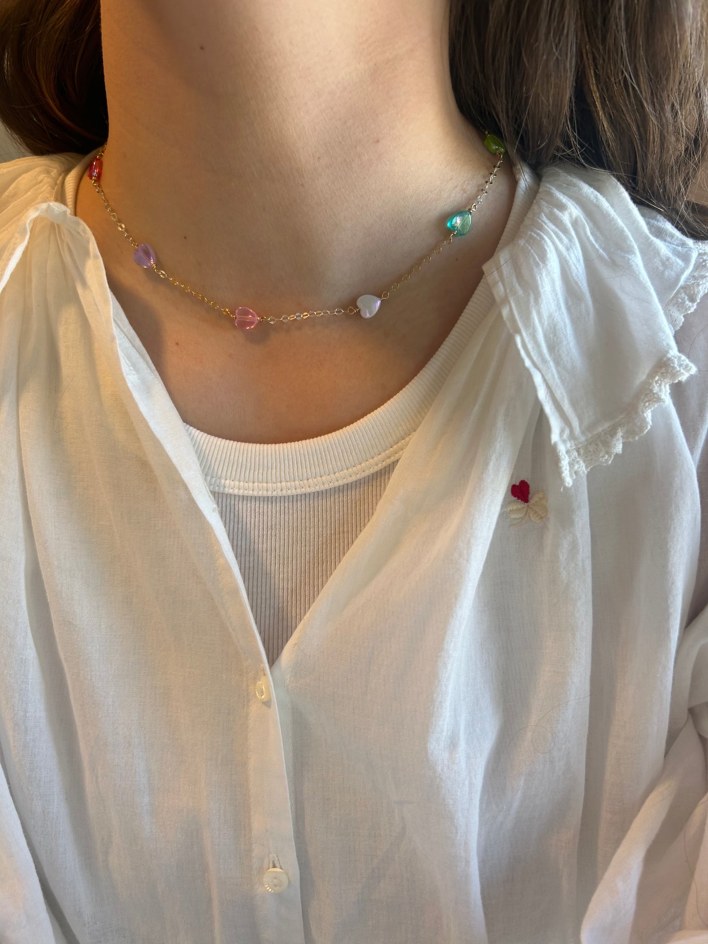 A person wears a white collared shirt with ruffled edges and a small red heart on the chest, accessorized with the Valentine Acrylic Heart Layer—a delicate 14k gold filled necklace featuring colorful heart beads and freshwater pearls.