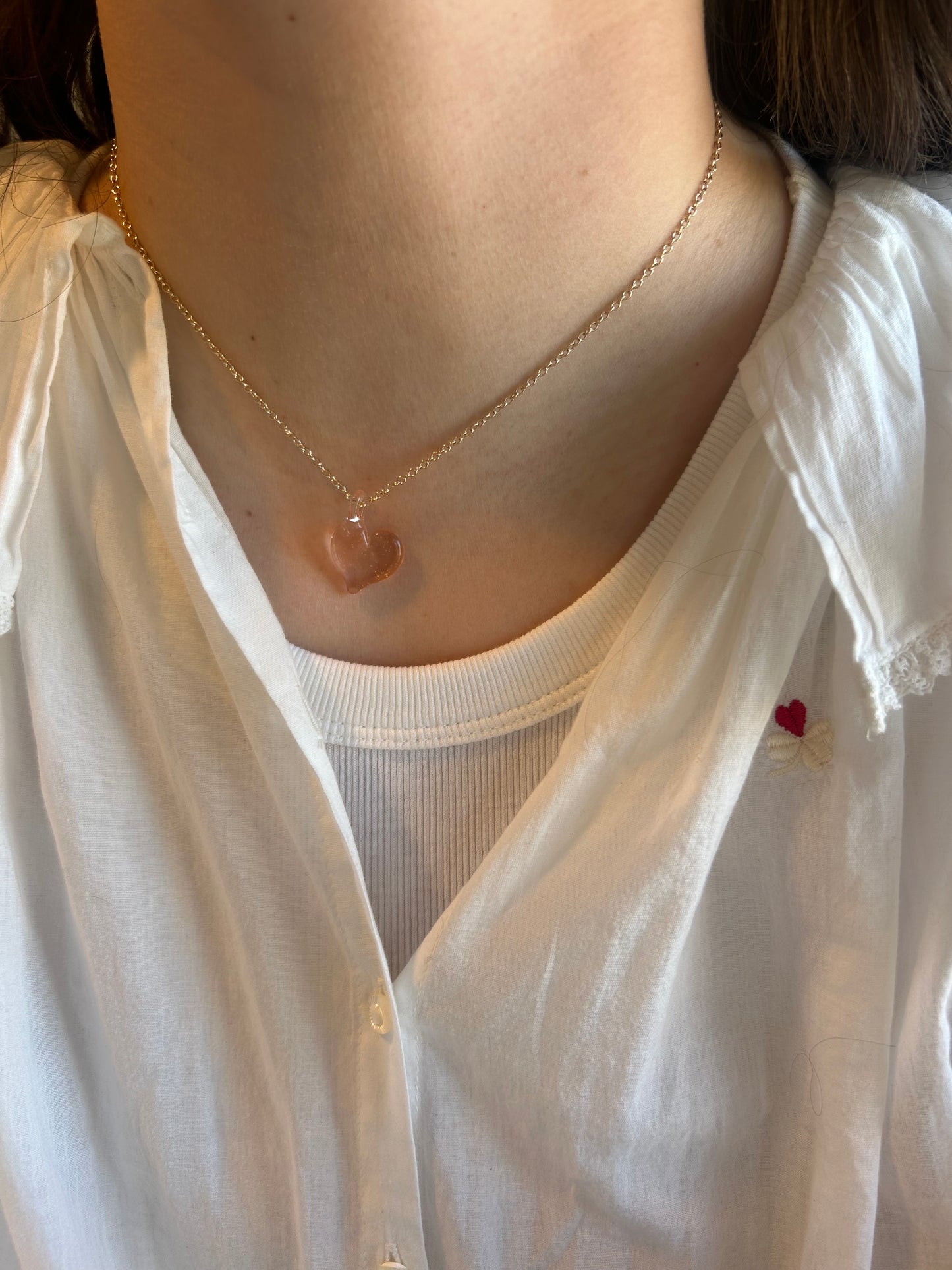 Someone wearing the Flat Glass Heart Layer—a delicate 14k gold-filled chain necklace with a transparent handblown glass heart-shaped pendant—styled with a white button-up shirt over a ribbed top featuring a small red embroidered heart.
