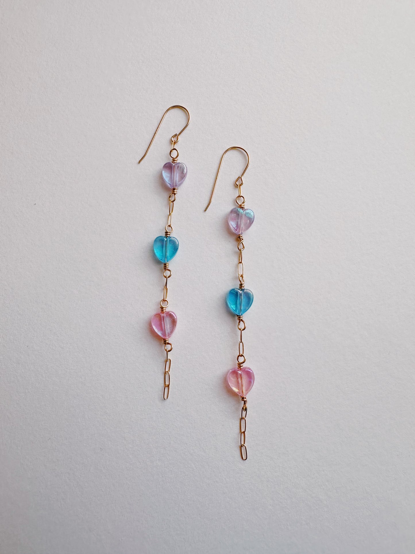 Acrylic Heart Drops: Dangling earrings with gold filled hooks and chain links, each showcasing three vertically arranged pastel acrylic heart beads in purple, blue, and pink on a plain white background.