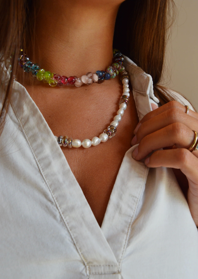 A woman in a white collared shirt wears the Glass Donut and Pearl Layer necklace, featuring colorful glass beads, freshwater pearls, and silver accents. She holds her collar with her left hand adorned with gold rings.