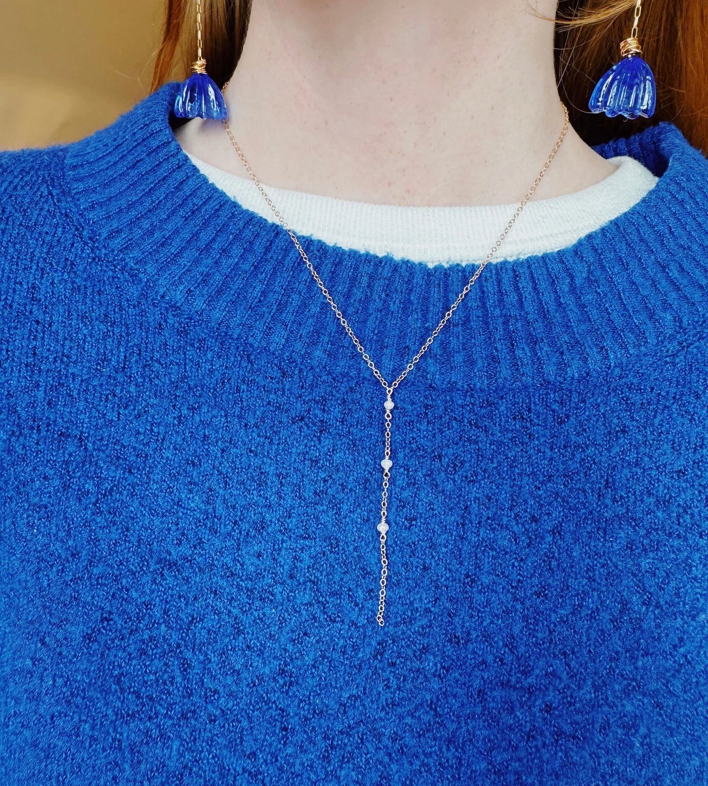 A person in a royal blue textured sweater and white undershirt wears the Dainty Pearl Lariat necklace and blue flower-shaped earrings. The jewelry, especially the delicate beaded lariat, stands out against the sweater.