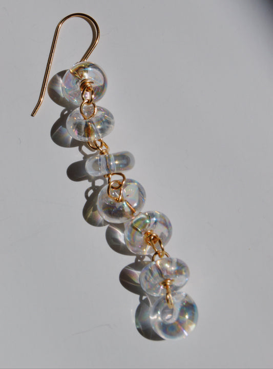 Donut Drops: Gold hook earrings featuring six clear, round handmade lampwork glass beads linked with gold, displayed on a white surface casting soft, colorful shadows—a striking handcrafted jewelry piece.