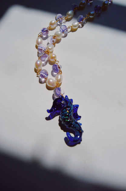 The Glass Seahorse Layer necklace features alternating cultured freshwater pearls and purple beads with a dark blue glass seahorse pendant, displayed on a white surface in gentle sunlight.