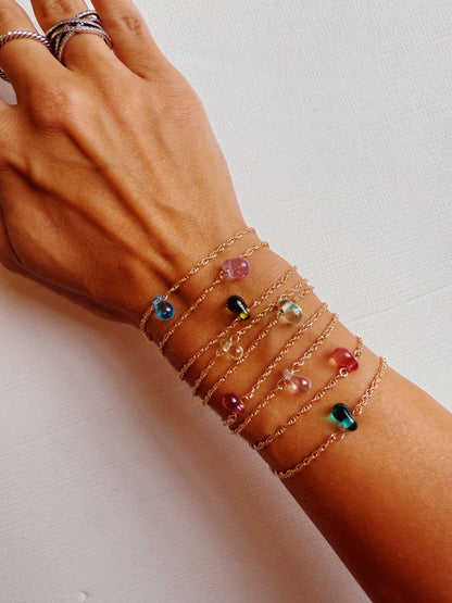 A hand showcases layered Dewdrop Bracelets—dainty, adjustable 14k gold filled chains with colorful glass beads—against a white textured background, highlighting their vibrant mix of stones.