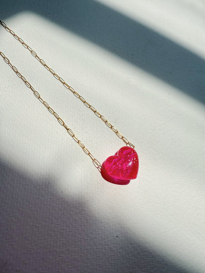 The Acrylic Heart Necklace, featuring a pink acrylic heart pendant, rests on a white textured surface bathed in soft, angled sunlight and gentle shadows.