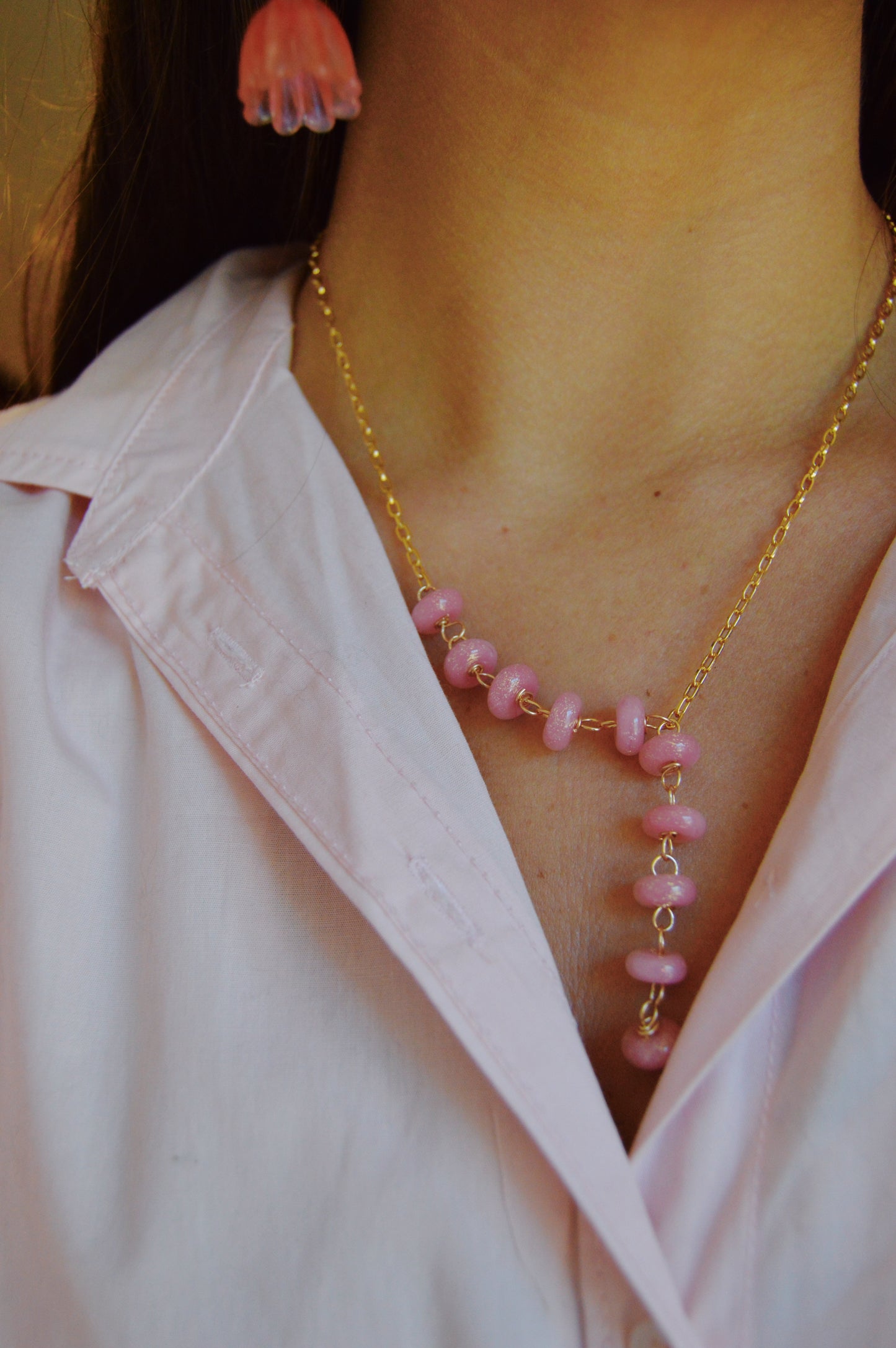 A person wears a light pink collared shirt, pink flower earring, and the Donut Lariat-Pink Sparkle necklace with pink beads and gold chain as the main focal point on their chest.