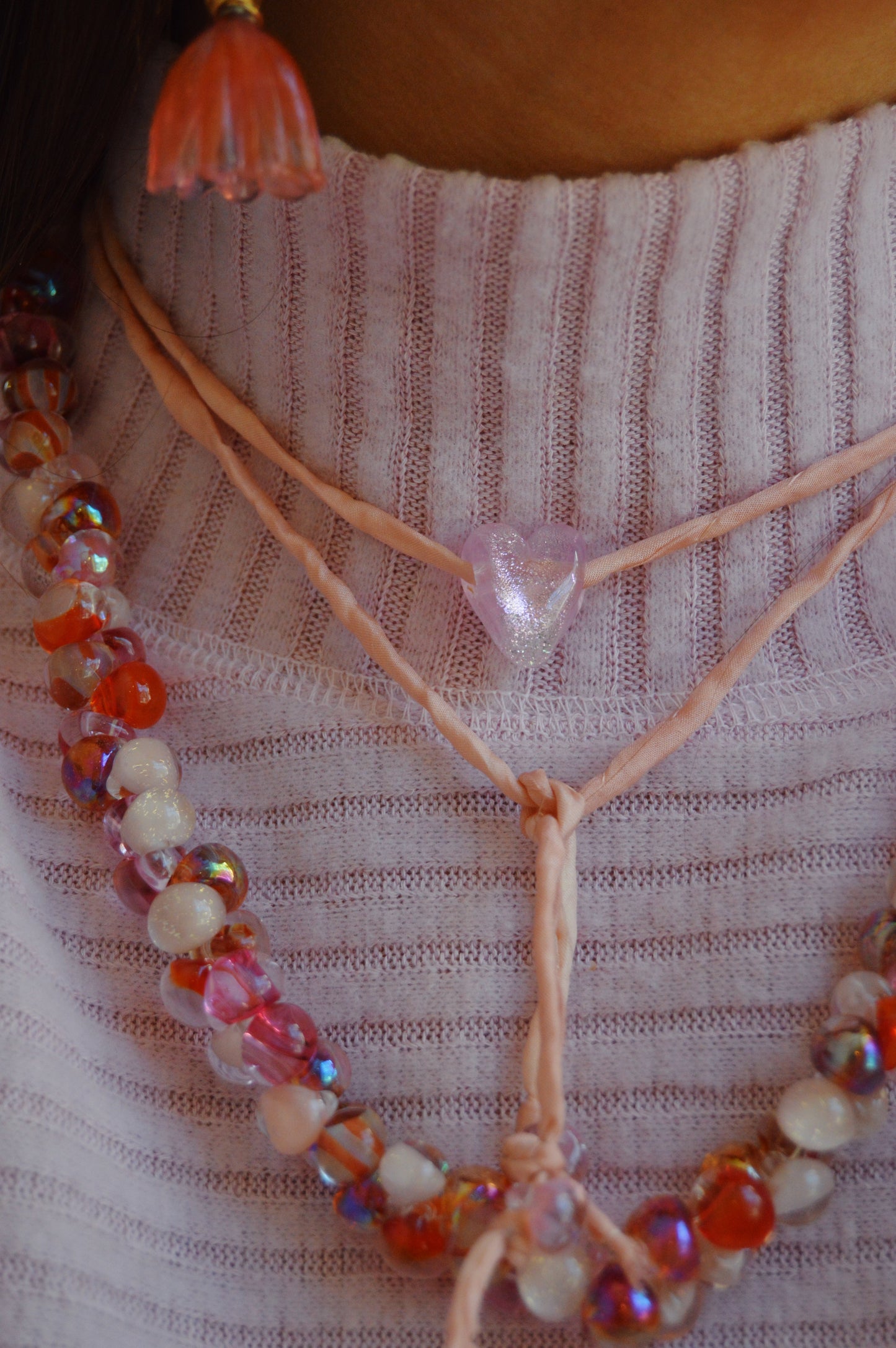 Close-up of a person wearing a pink knit top, the Valentine Bubble Glass Necklace- Lovebug featuring pink and white lampwork beads, a pink heart-shaped pendant necklace, and a pink flower-shaped earring.