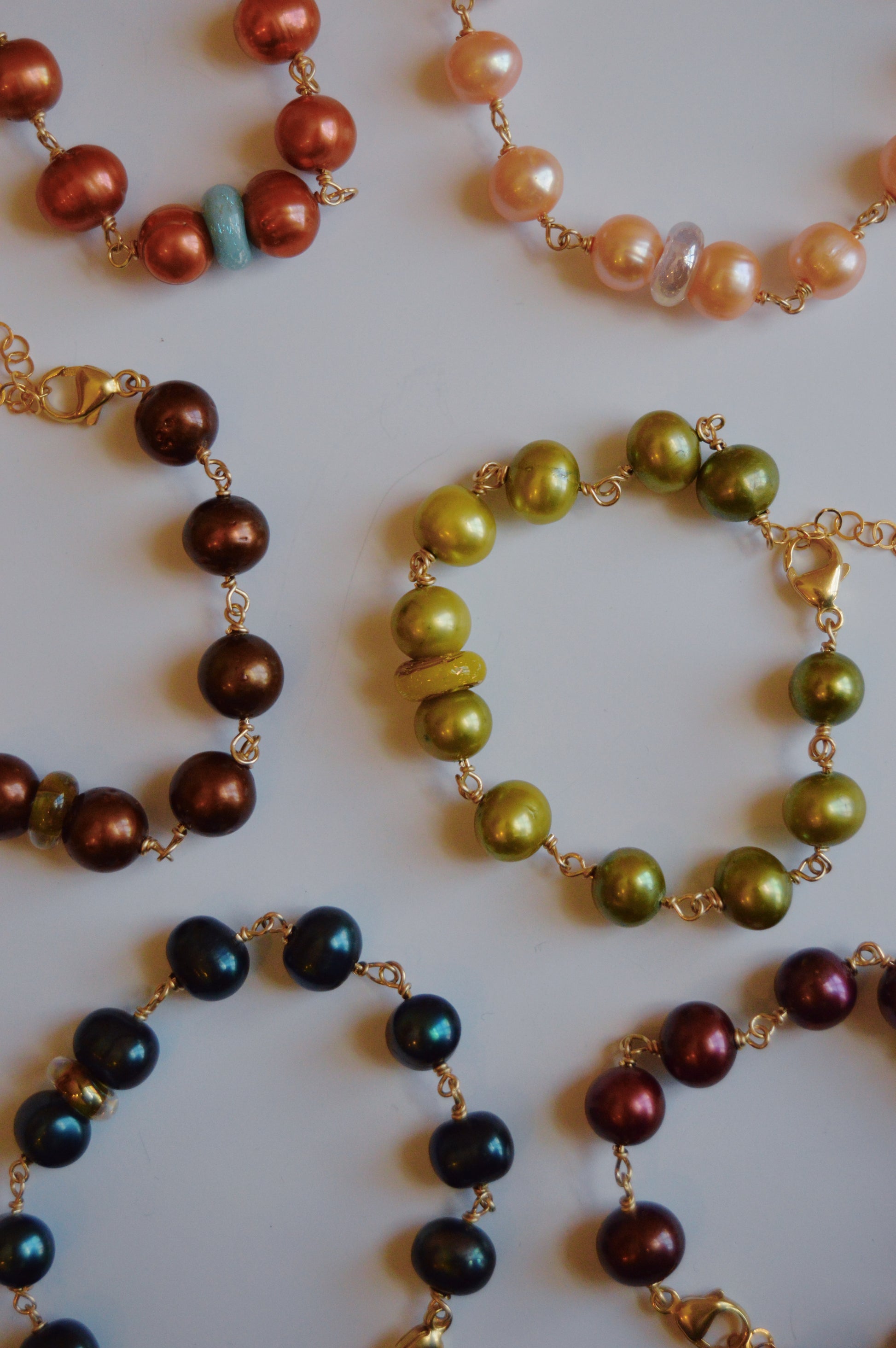 The Pearl and Glass Bracelet features shiny, colorful cultured freshwater pearls in brown, green, blue, red, and peach, arranged in a scattered pattern on a white background.
