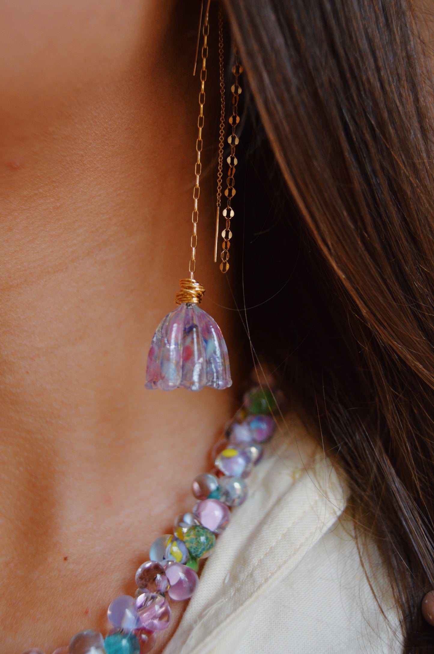 Close-up of a woman's neck and shoulder wearing the Glass Tulips Earrings- Bloom: a long oxidized sterling silver chain earring with a purple-blue glass flower pendant, alongside a colorful beaded necklace and her brown hair partially visible.