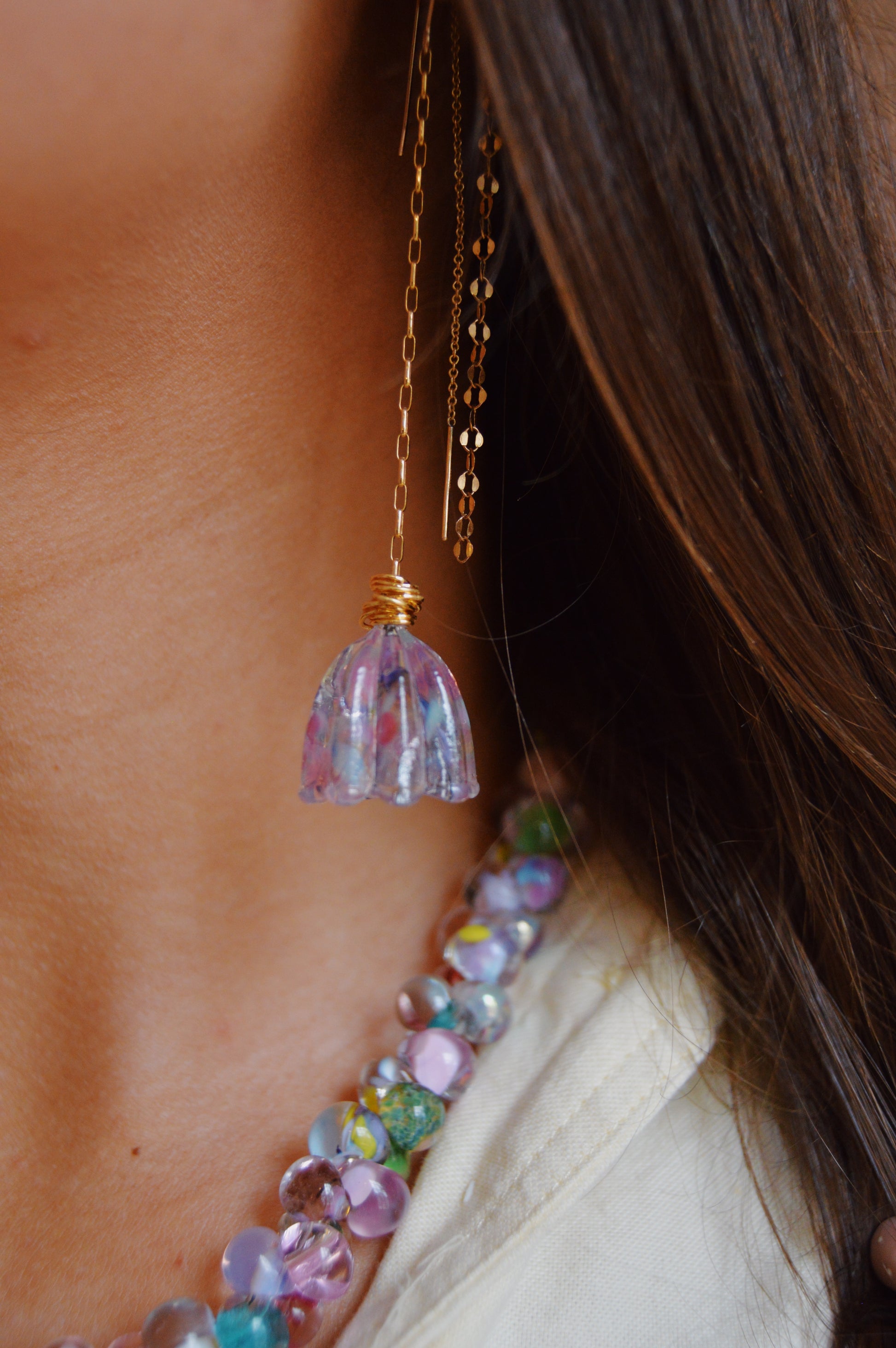 Close-up of a woman's neck and shoulder wearing the Glass Tulips Earrings- Bloom: a long oxidized sterling silver chain earring with a purple-blue glass flower pendant, alongside a colorful beaded necklace and her brown hair partially visible.