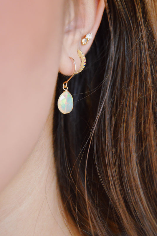 Close-up of a person's ear with three earrings: a small 14k gold-filled stud, a gold hoop with a leaf design, and the Baby Opal Drops—an opal earring featuring a teardrop-shaped pendant. Brown hair is partially visible in the background.