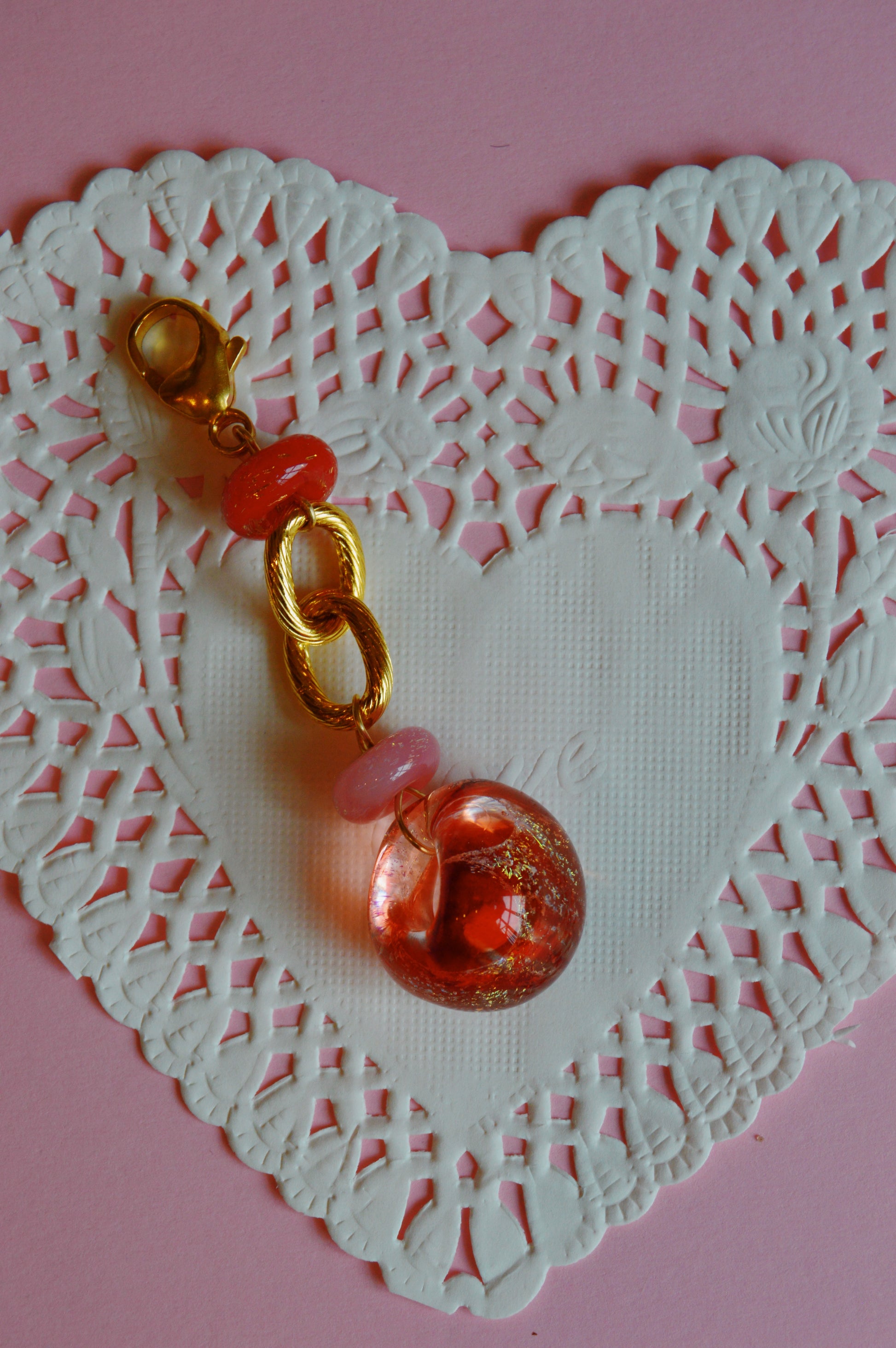 The Valentine Grand Bubble Charm, a gold keychain with pink and red beads and a round glass pendant, rests on a heart-shaped white lace paper doily against a pink background.
