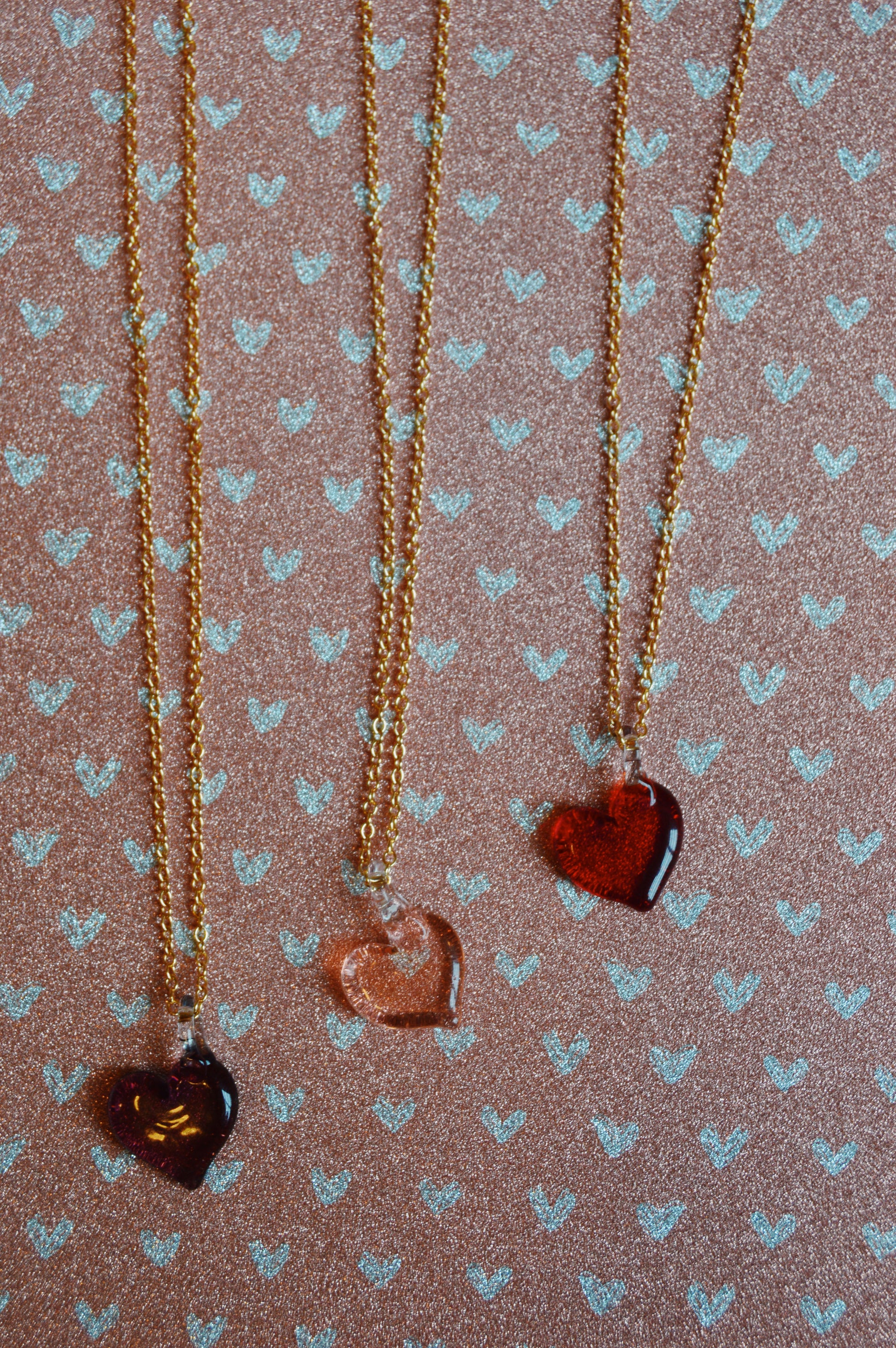 Three Flat Glass Heart Layer necklaces—brown, clear, and red—hang on gold-filled chains, displayed on a pink surface with white heart patterns.