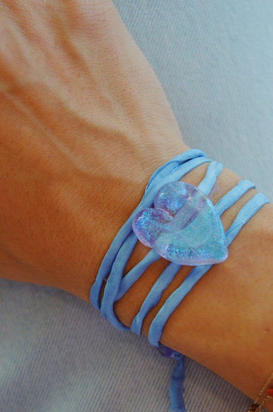 A hand wears the Silk Glass Heart- Periwinkle bracelet, featuring a light blue silk wrap and a shiny, translucent periwinkle lampwork glass heart charm, resting on a matching light blue fabric background.