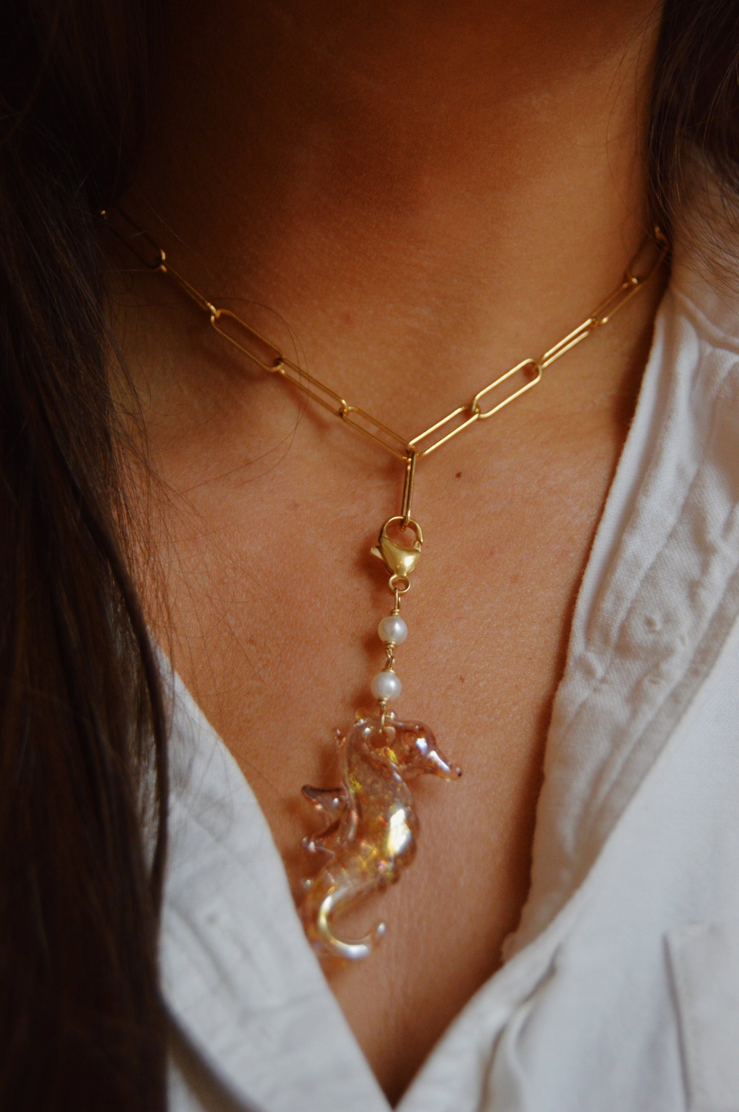A person wearing a white shirt and the Seahorse Charm—a 14k gold-filled chain necklace with a glass seahorse pendant and small freshwater pearls above it.