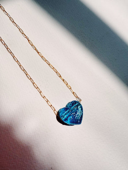 The Acrylic Heart Necklace, featuring a blue heart pendant on a gold filled chain, is displayed on a white textured surface with sunlight and shadows highlighting its details.