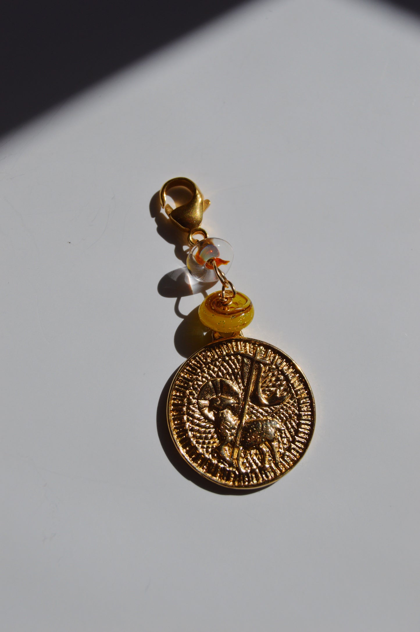 The Lamb of God Charm features a gold coin-shaped pendant with an engraved figure and a handmade lampwork glass bead, attached to a keychain clasp with 14k gold-filled findings, displayed on a white surface with a shadow in the corner.