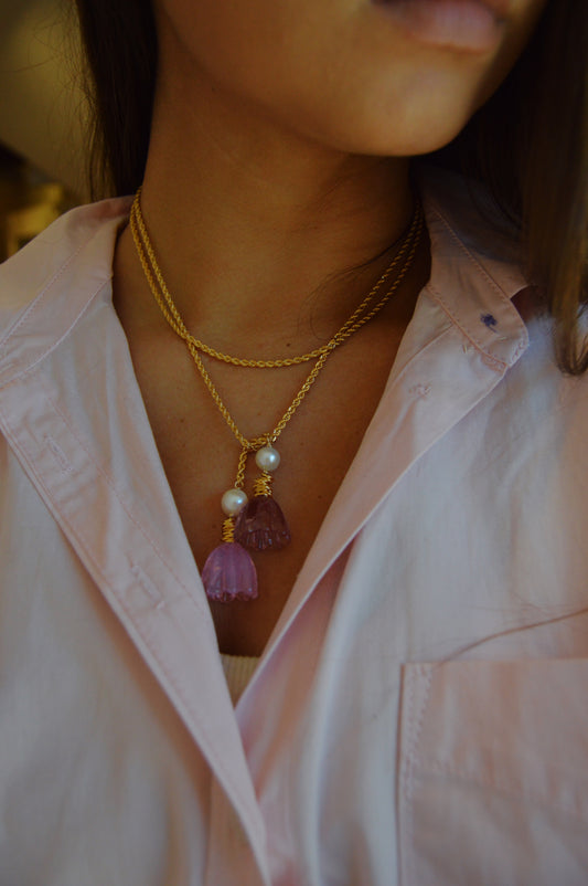 A woman models the Tulip Wrap Necklace-Ballet Slippers, featuring gold chains with pearl and purple stone pendants, paired with a pale pink collared shirt. The image is cropped to highlight her neck and part of her face.