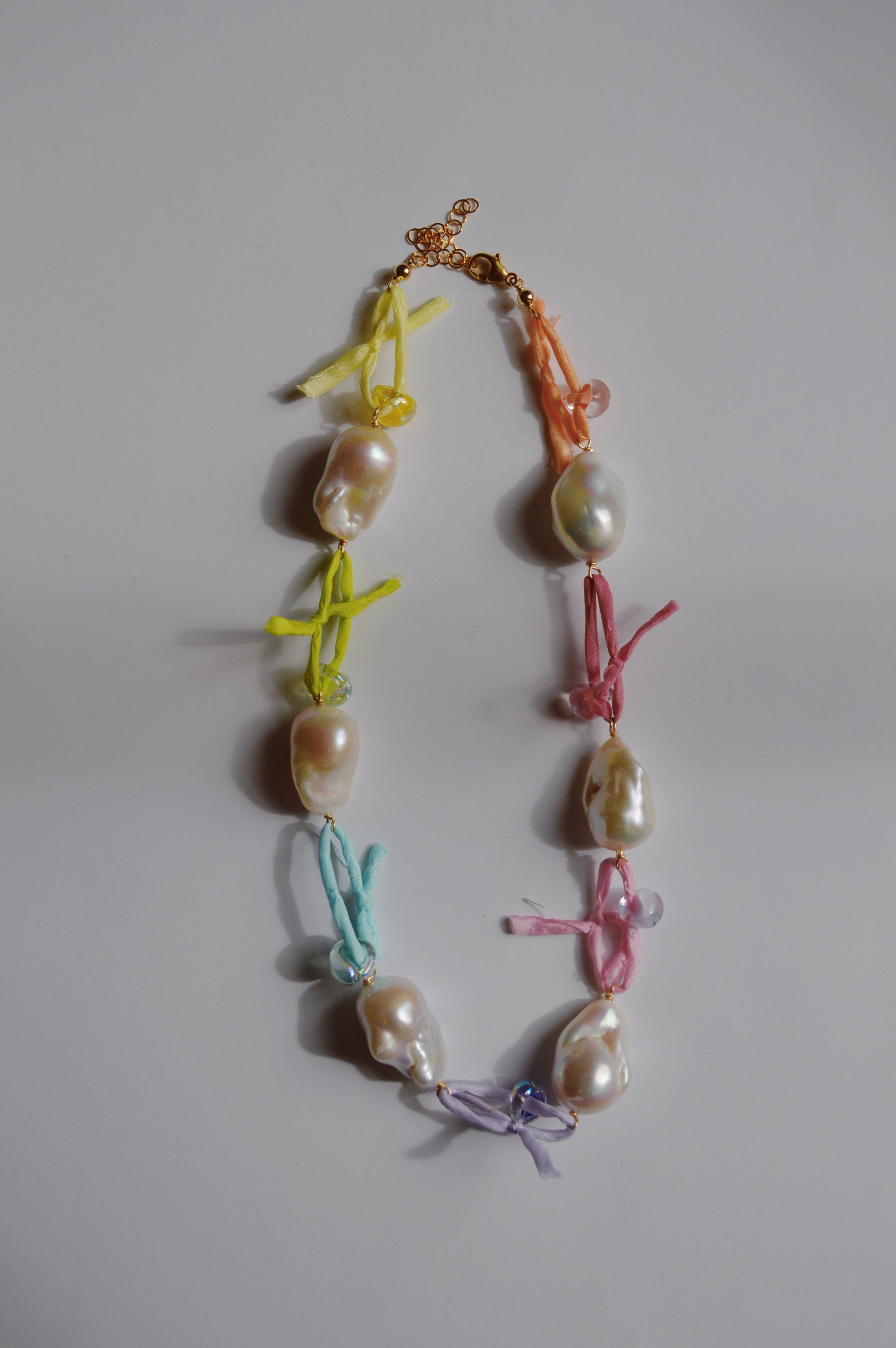 The Silk and Pearl Layer- Rainbow necklace features large, irregular freshwater pearls separated by colorful fabric knots in yellow, blue, purple, pink, and orange, finished with a gold clasp and shown on a white background.