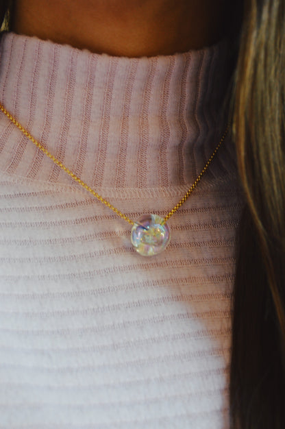 A close-up of a person wearing a ribbed light pink turtleneck sweater and the Grand Bubble Single - Constellation, a delicate gold filled chain necklace featuring a round, iridescent glass pendant.